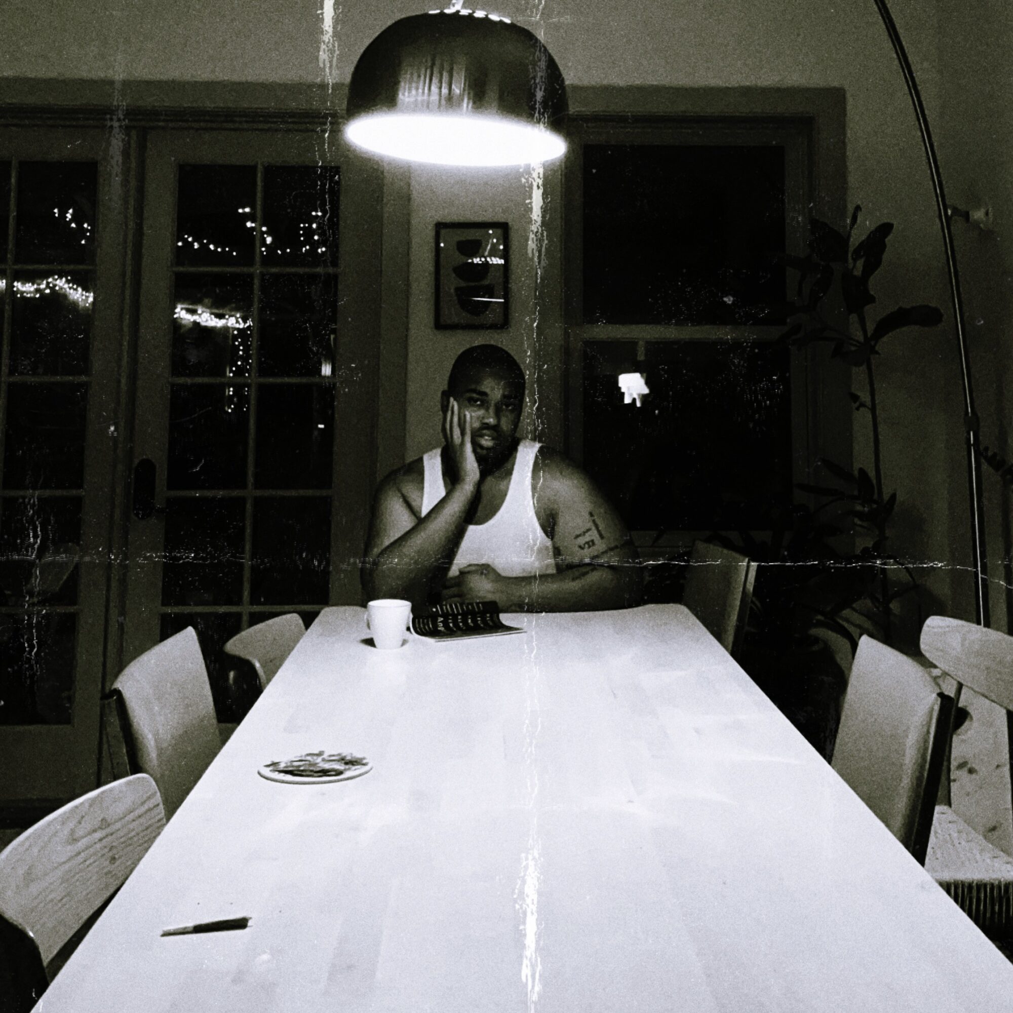 Person sitting at a long dining table with a cup, phone, and plates, illuminated by a hanging light, in a dark room.