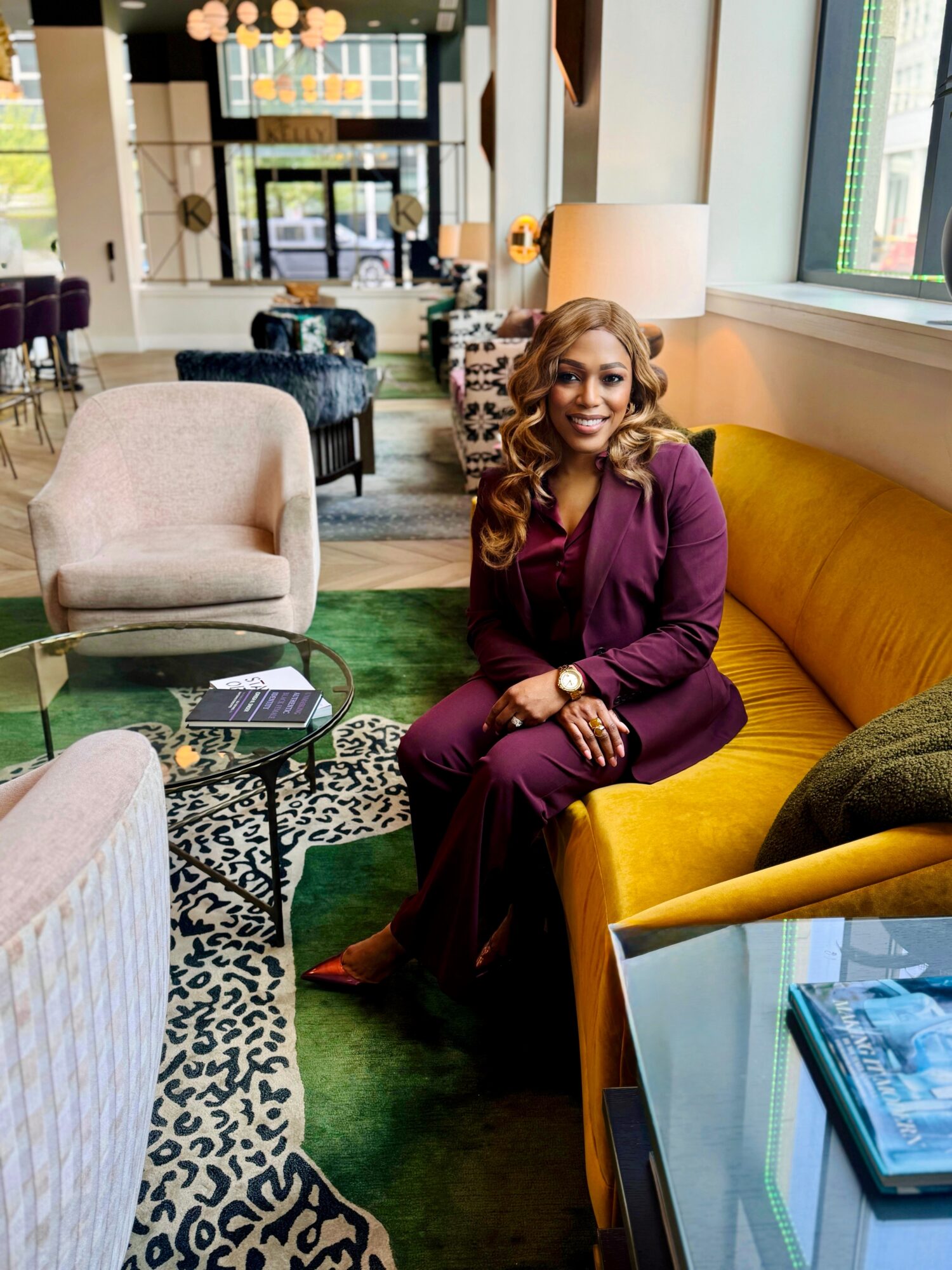 Woman with long hair in a purple outfit sitting on a yellow sofa in a modern, well-lit lounge area with large windows.