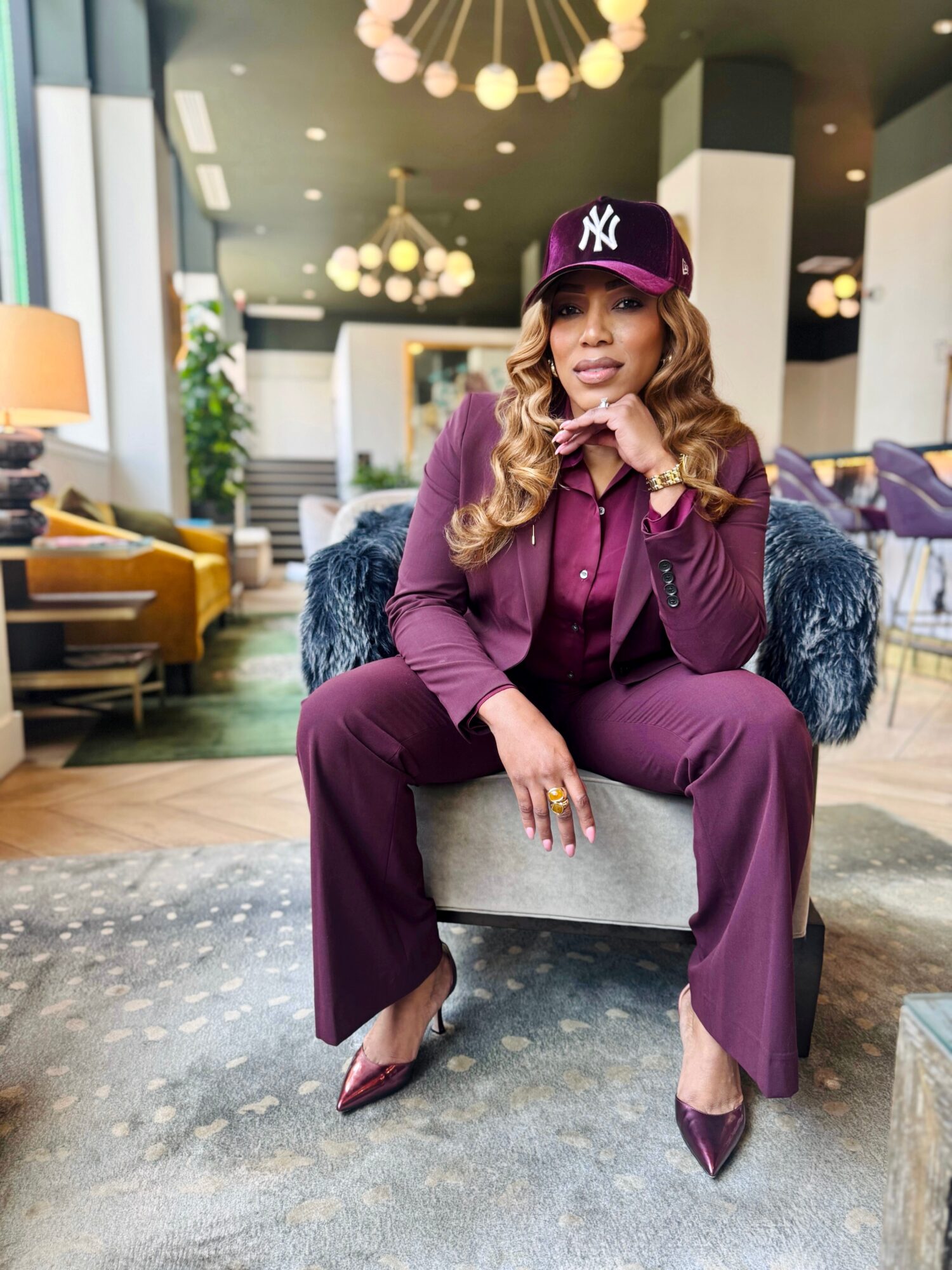 Woman in purple suit and cap sitting on a chair in a modern, well-lit lounge area with plants and furniture.