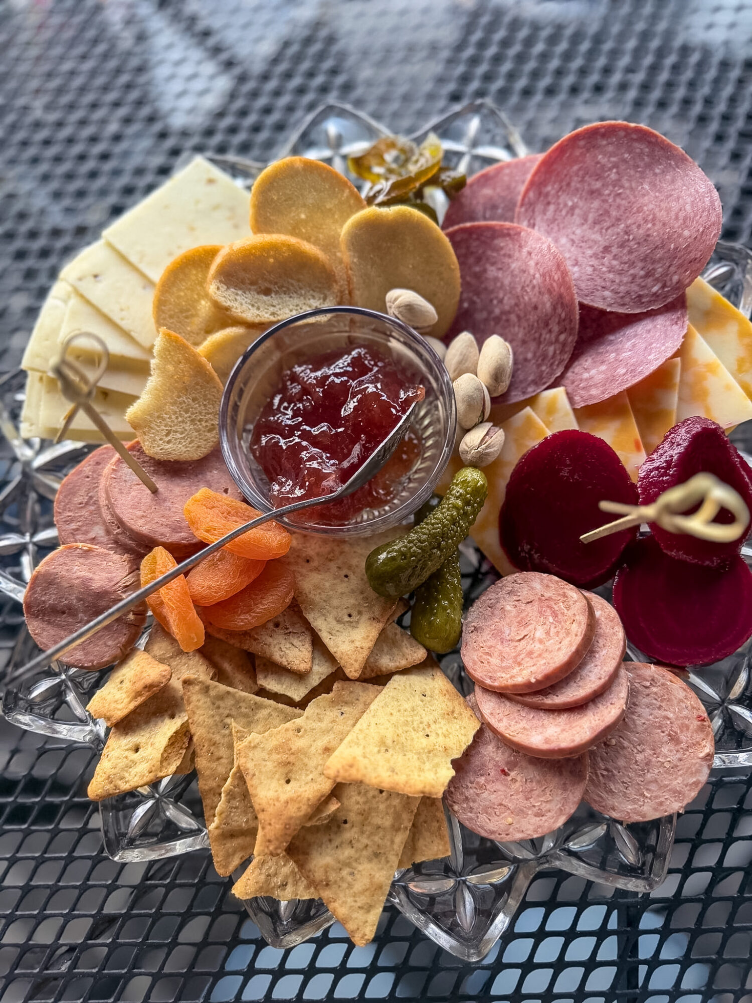 Assorted charcuterie with crackers, sliced meats, cheese, pickles, and a small bowl of jam on a glass platter.