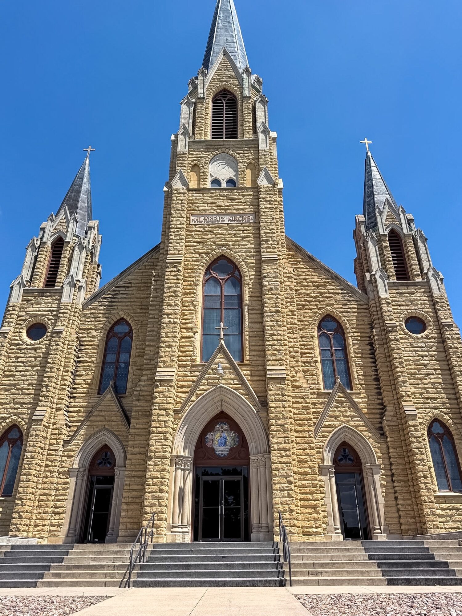 Front view of a church with three tall spires, arched windows, and stone steps leading to entrance, against a blue sky.