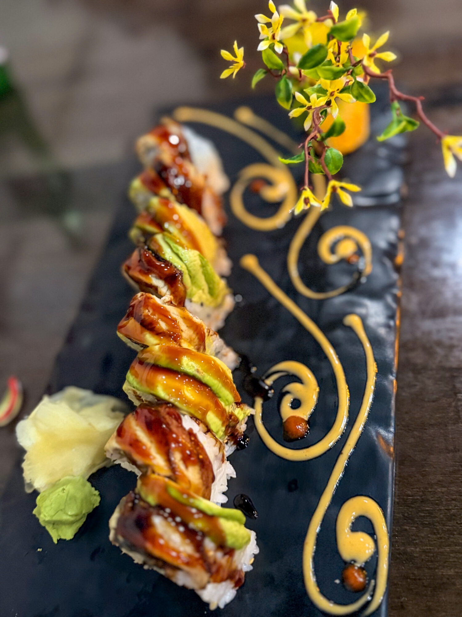 Sushi roll with avocado and fish, garnished with lettuce, on a decorative black plate with yellow swirl design and yellow flowers.