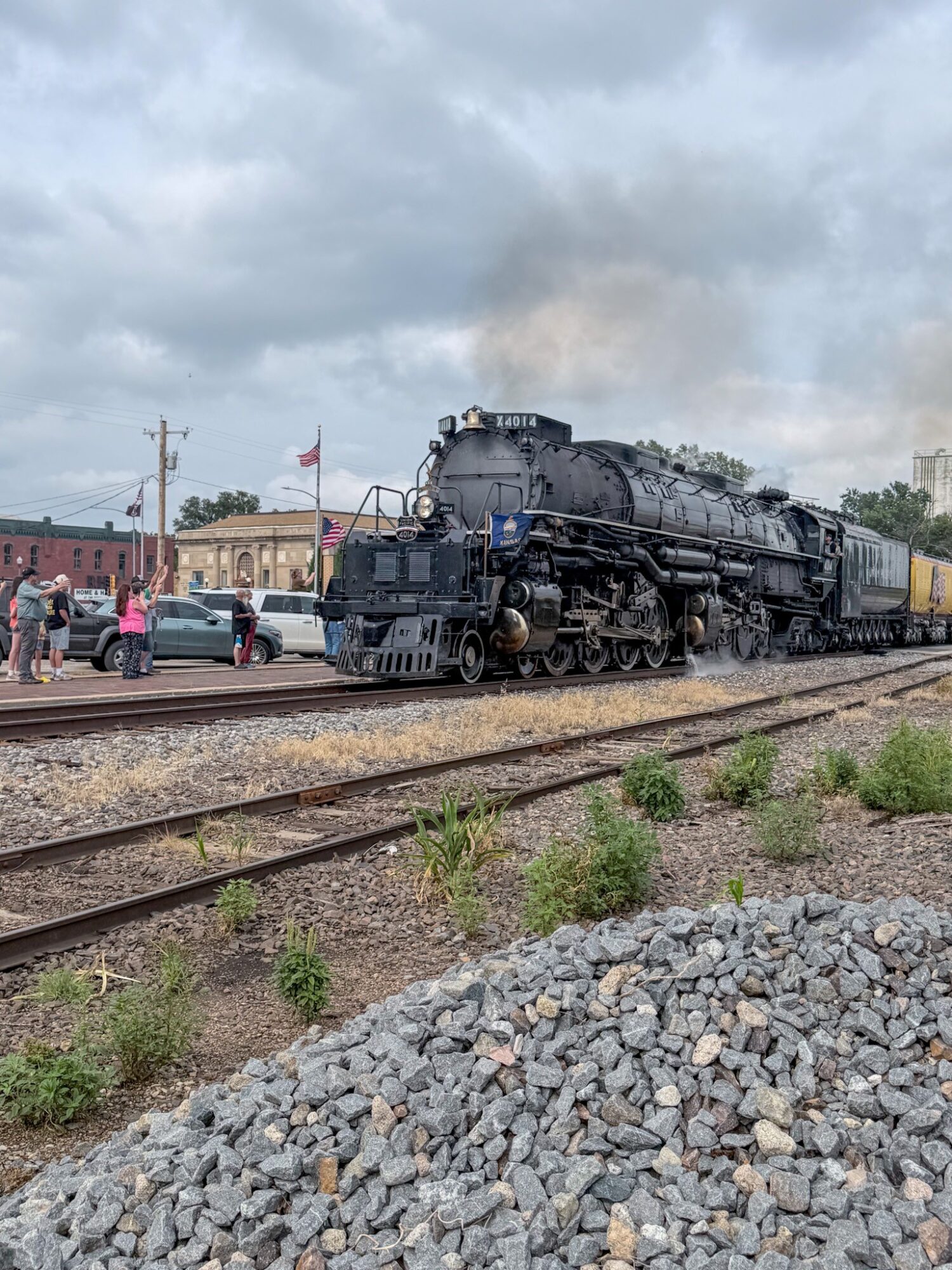 Steam locomotive on tracks with people and buildings in background under cloudy sky.