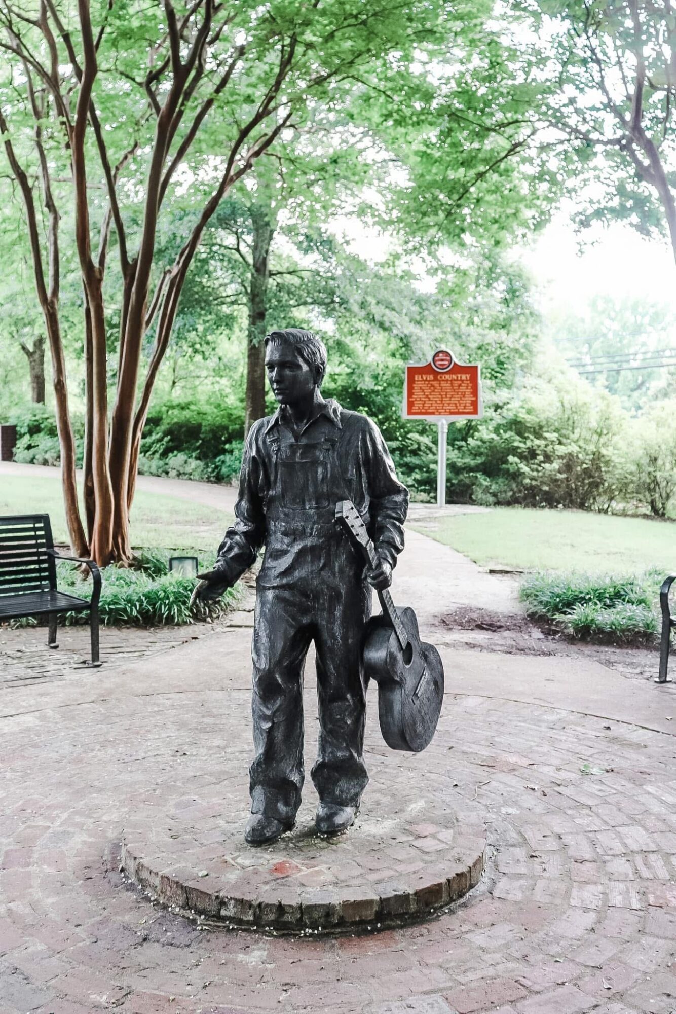 Bronze statue of a man holding a hat, standing on a circular stone platform in a park with trees and a bench.