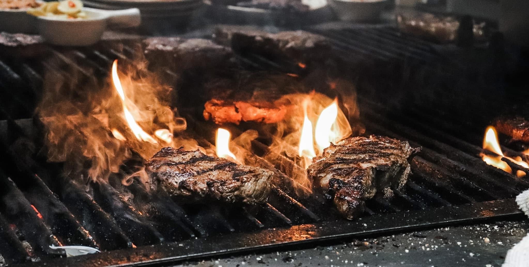 Meat grilling over open flames on a barbecue grill with visible flames and charred meat.