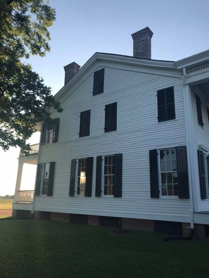 White house with black shutters, multiple windows, and a chimney, viewed from the side with a grassy yard and trees nearby.