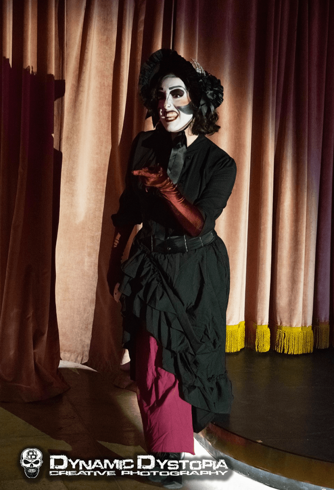 Person with makeup and costume standing on stage with curtains in background.