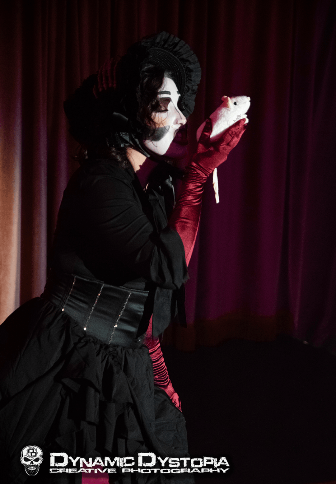 Person dressed as a mime with face paint, wearing a black outfit and red gloves, holding a small white object, standing in front of a curtain.