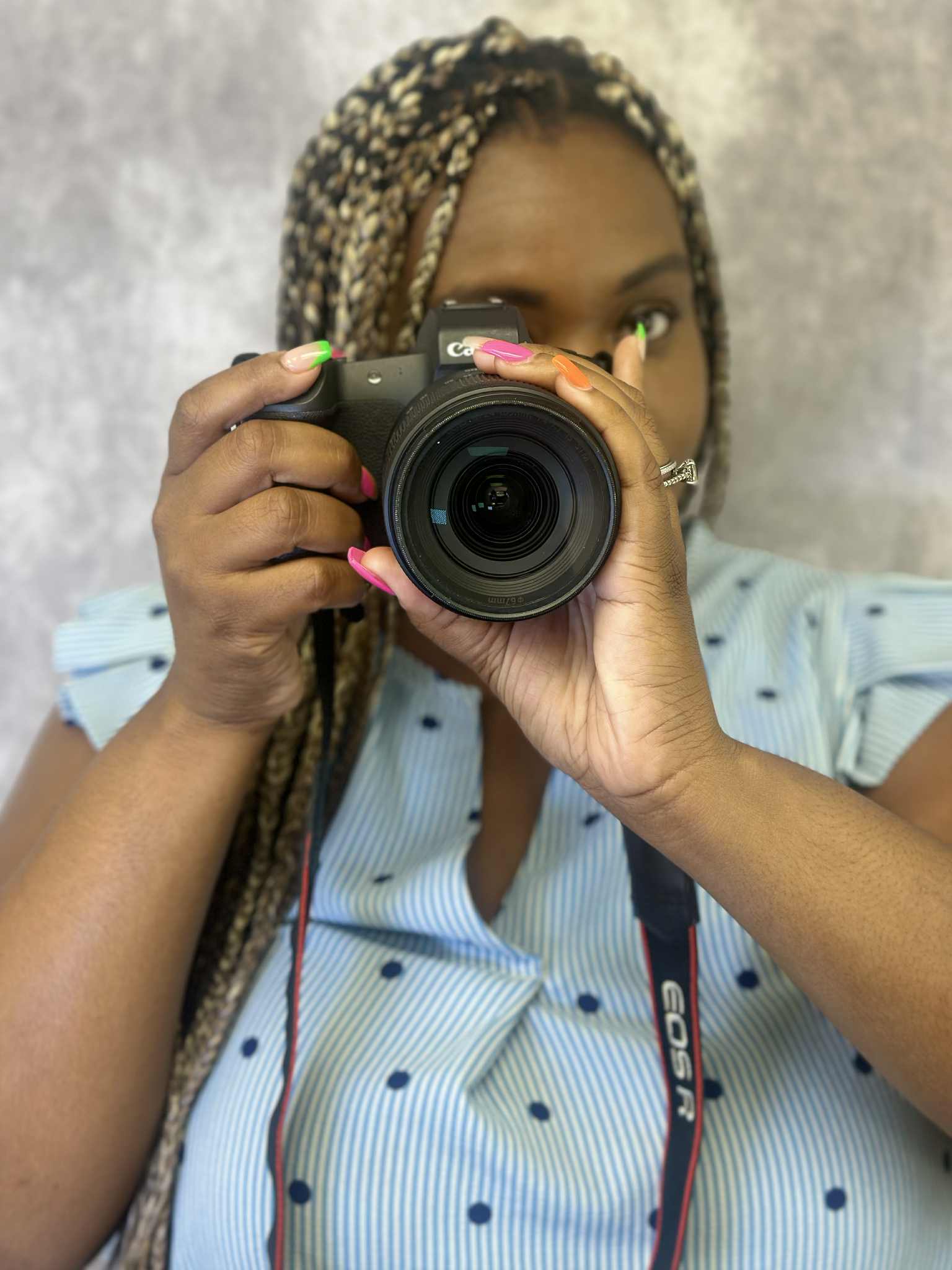 Person with braided hair taking a selfie with a camera, wearing a light blue shirt with polka dots, against a textured background.