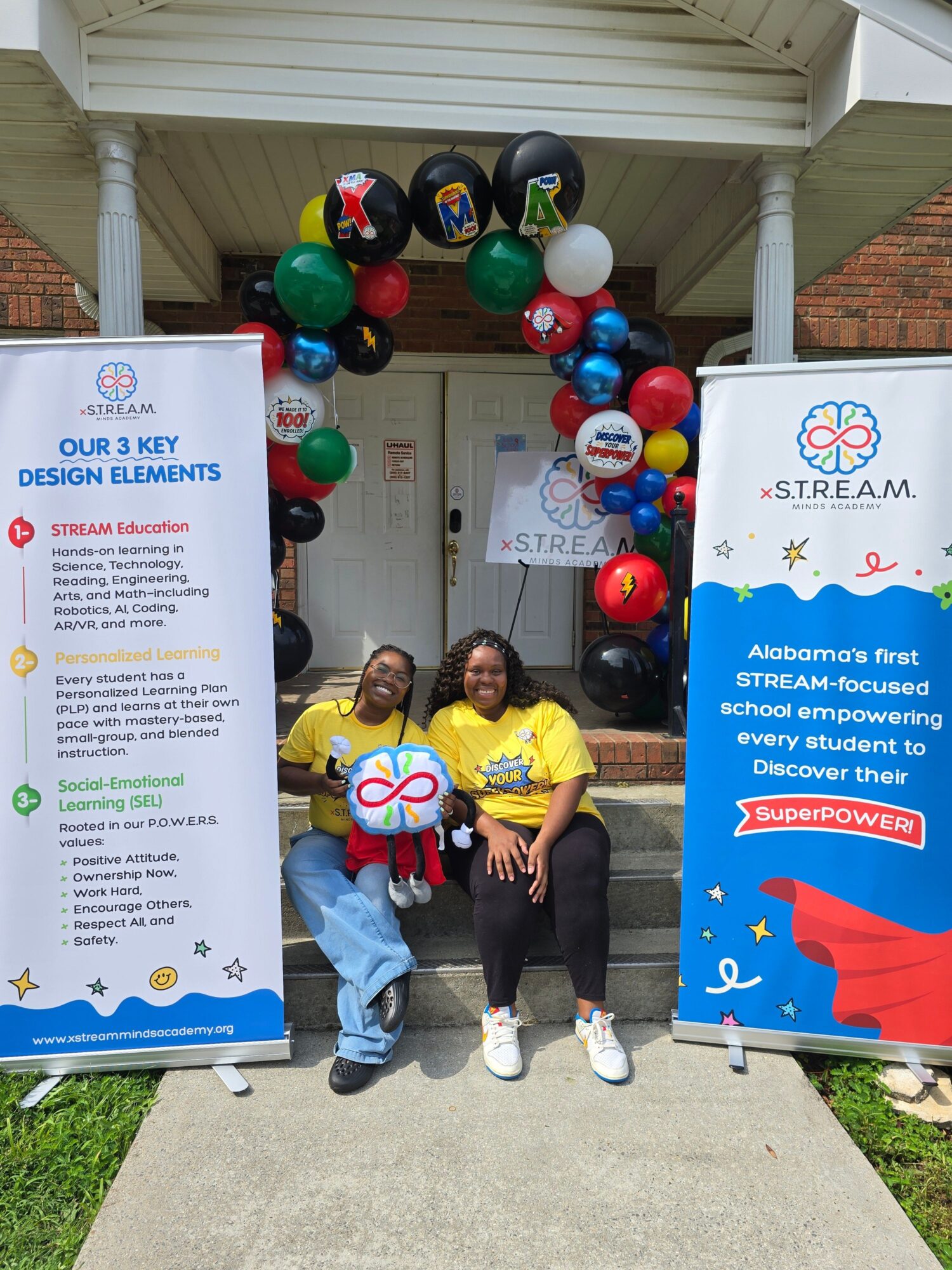 Three children stand outdoors near a display with balloons and banners, celebrating a special day or event.