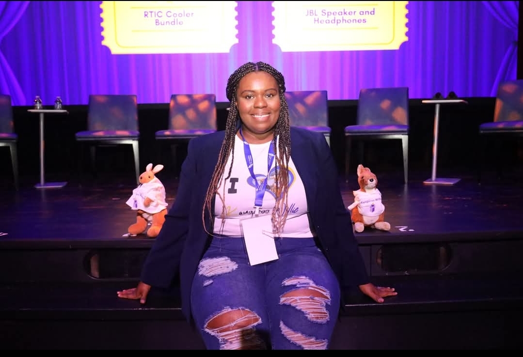 Woman sitting on stage with purple curtains, chairs, and two plush toys behind her, smiling at the camera.