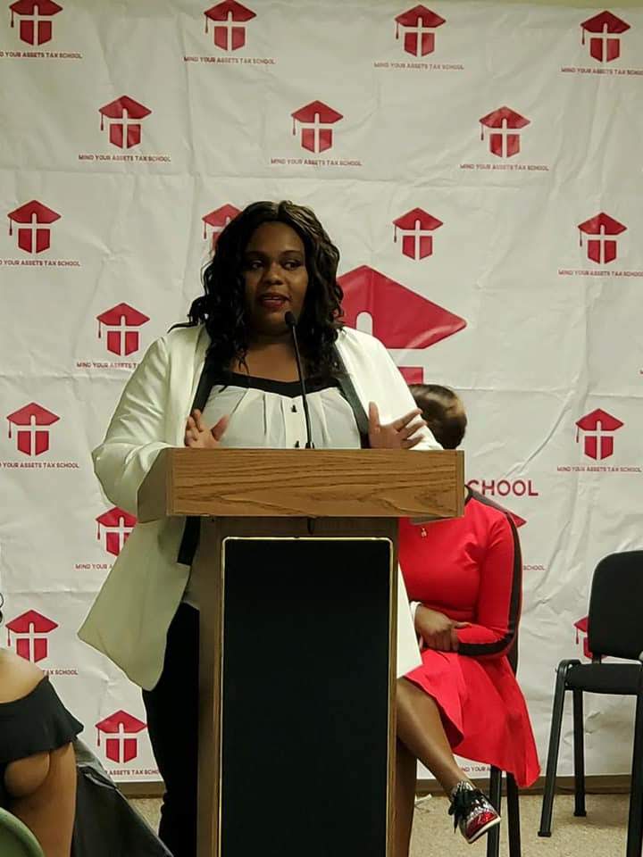 Woman speaking at a podium with a backdrop featuring a red and white logo, wearing a white blazer and black top.