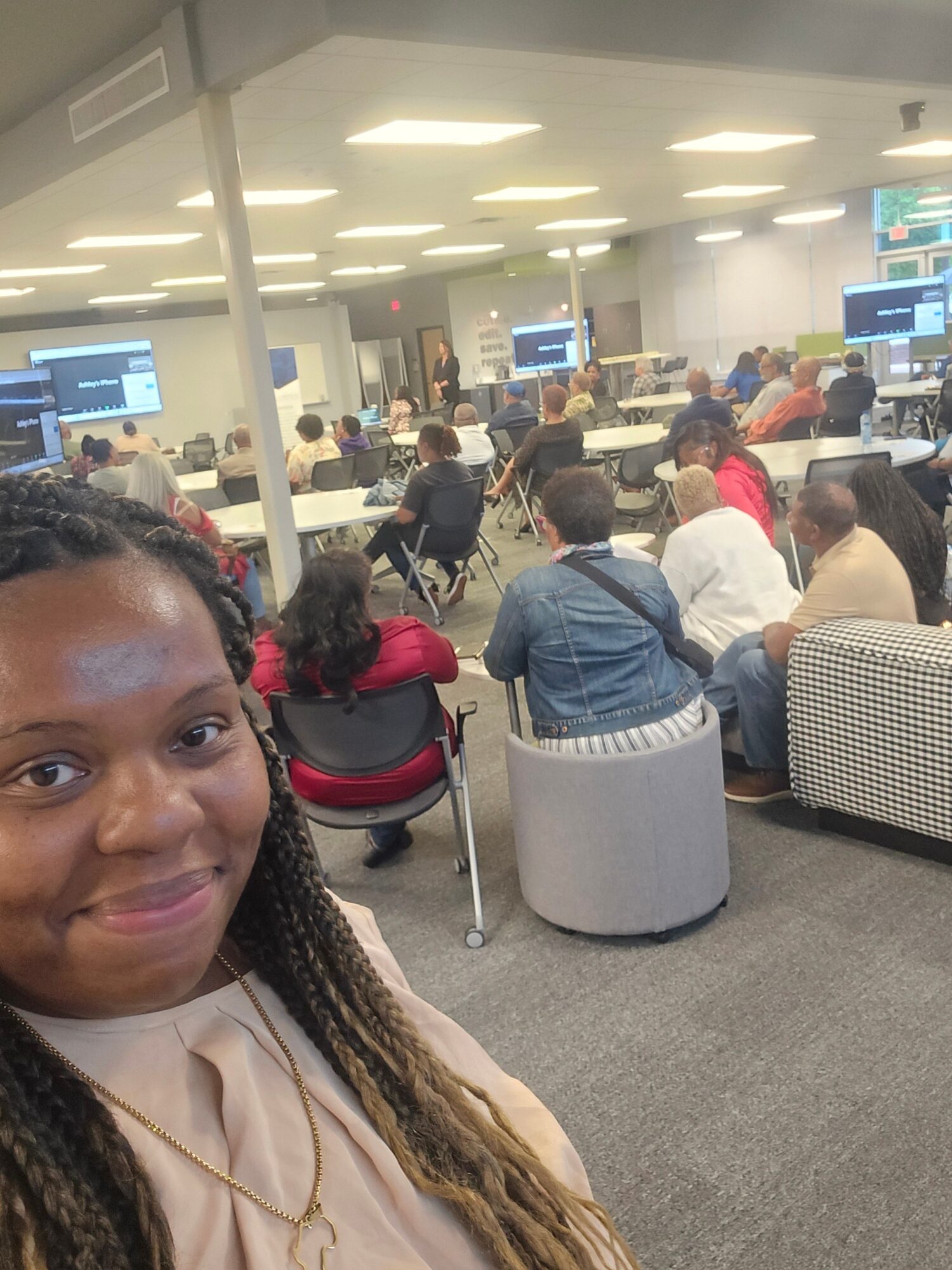 Woman with braided hair taking a selfie in a busy indoor setting with people and computer screens.