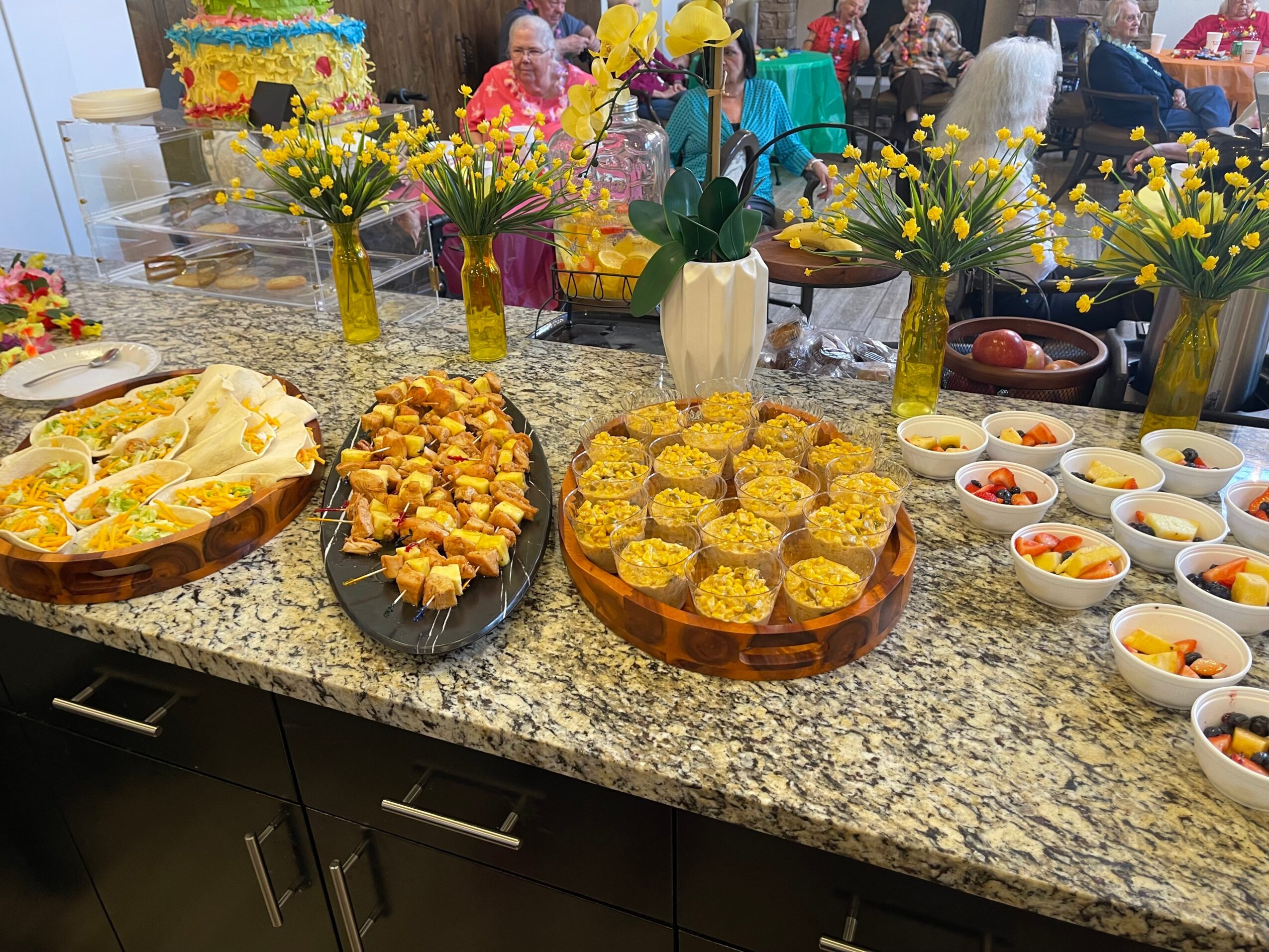 Display of food and flowers on a granite countertop with people in the background.