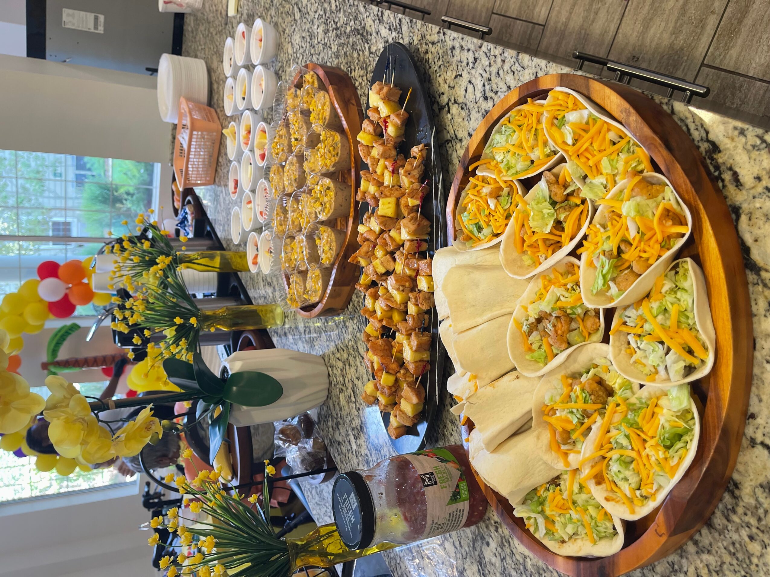 Table with tacos, chips, and various dishes, decorated with flowers and balloons, in a bright room with windows.
