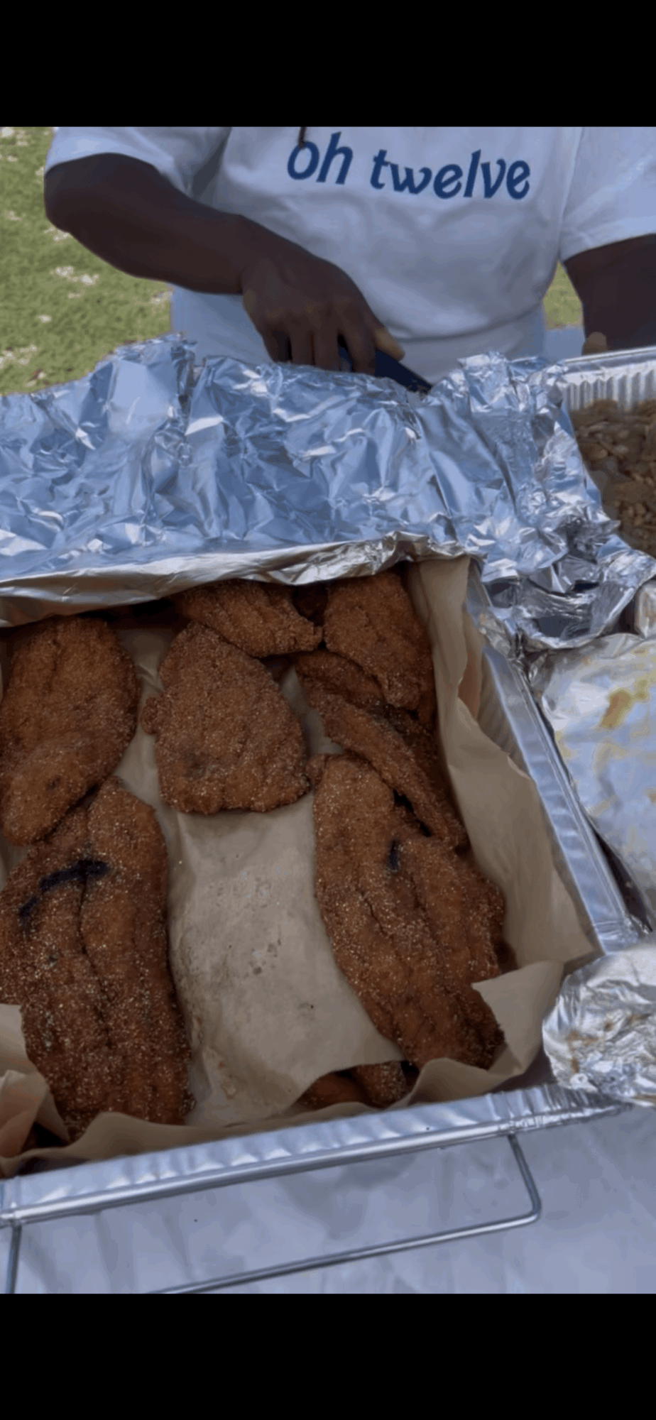 Person holding a tray of fried chicken and a sandwich with foil wrapping, outdoors on grass.