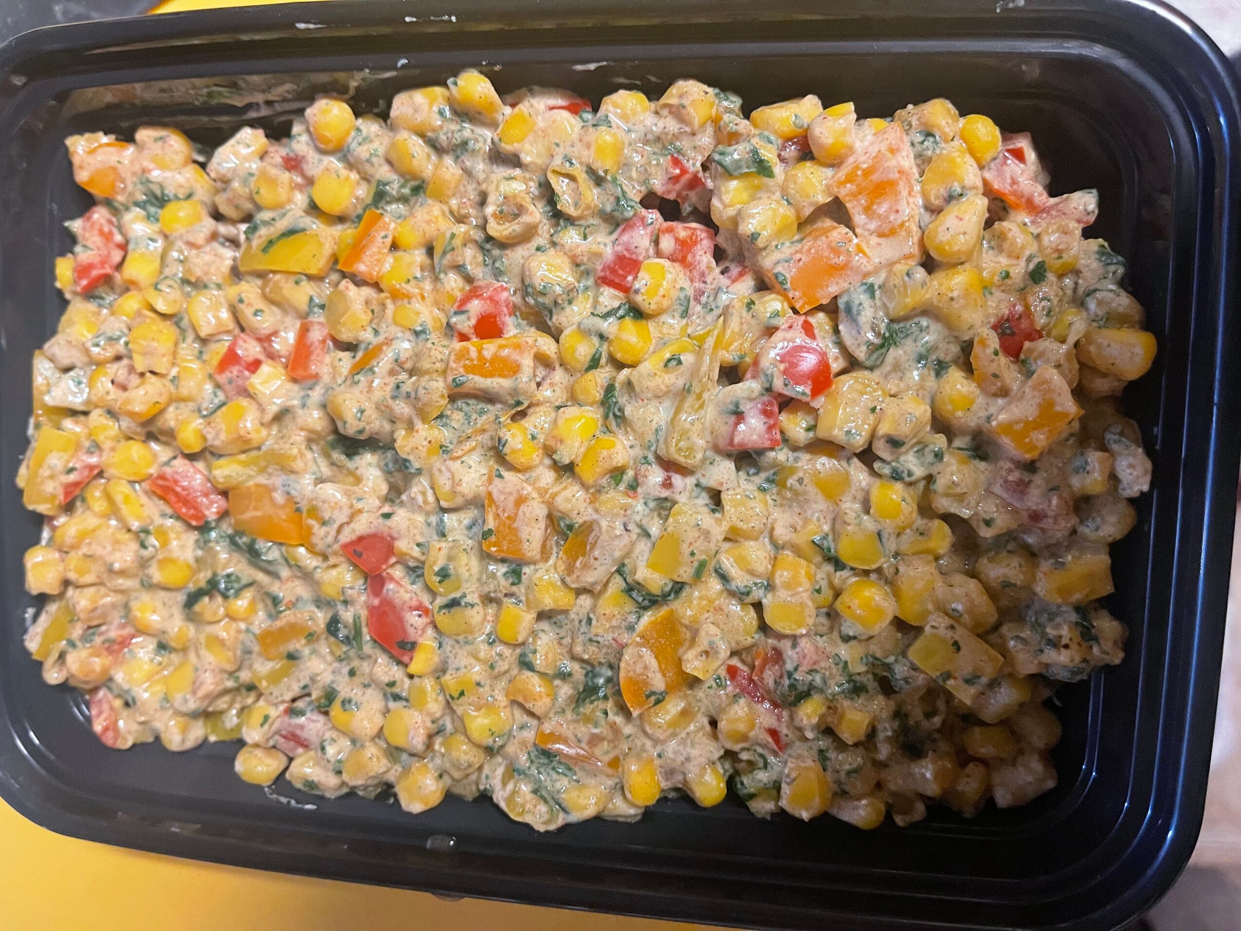 Colorful mixed vegetable salad in a black container, with visible corn, red peppers, and other vegetables, on a yellow surface.