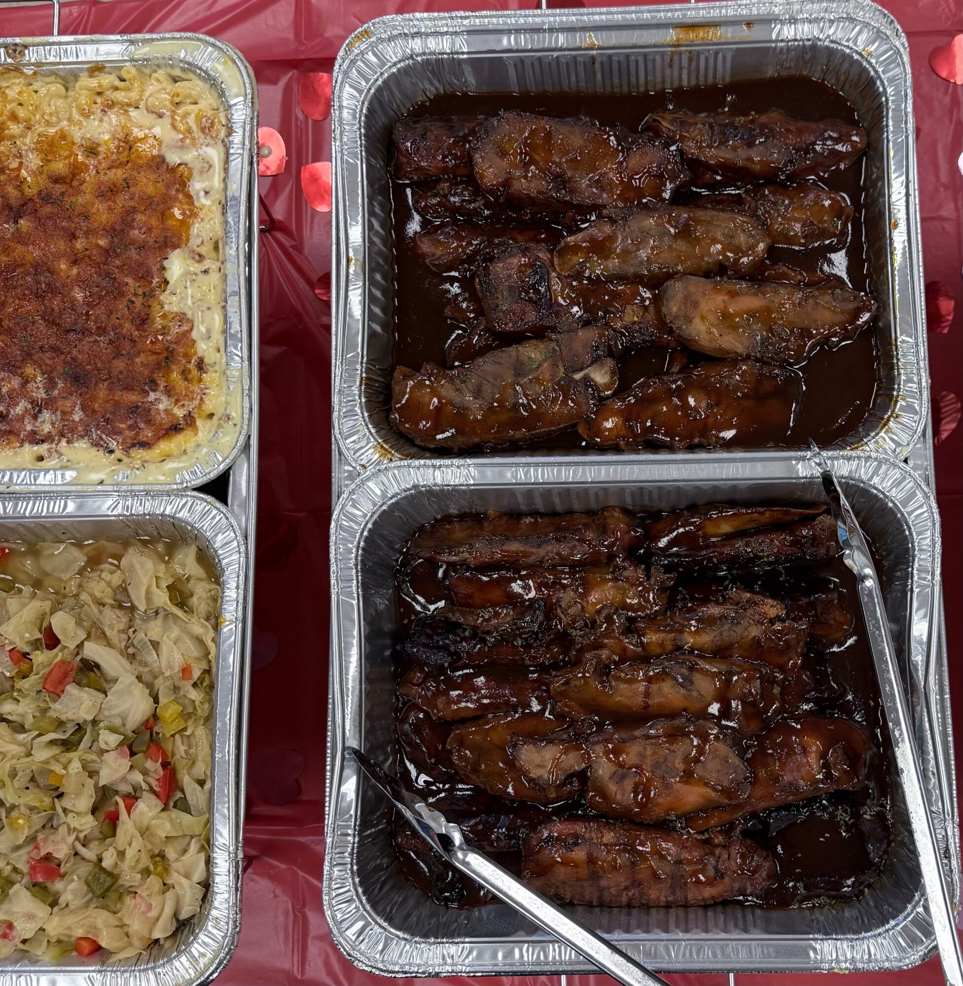 Two aluminum trays with glazed ribs and two trays with baked casserole dishes, on a red surface.