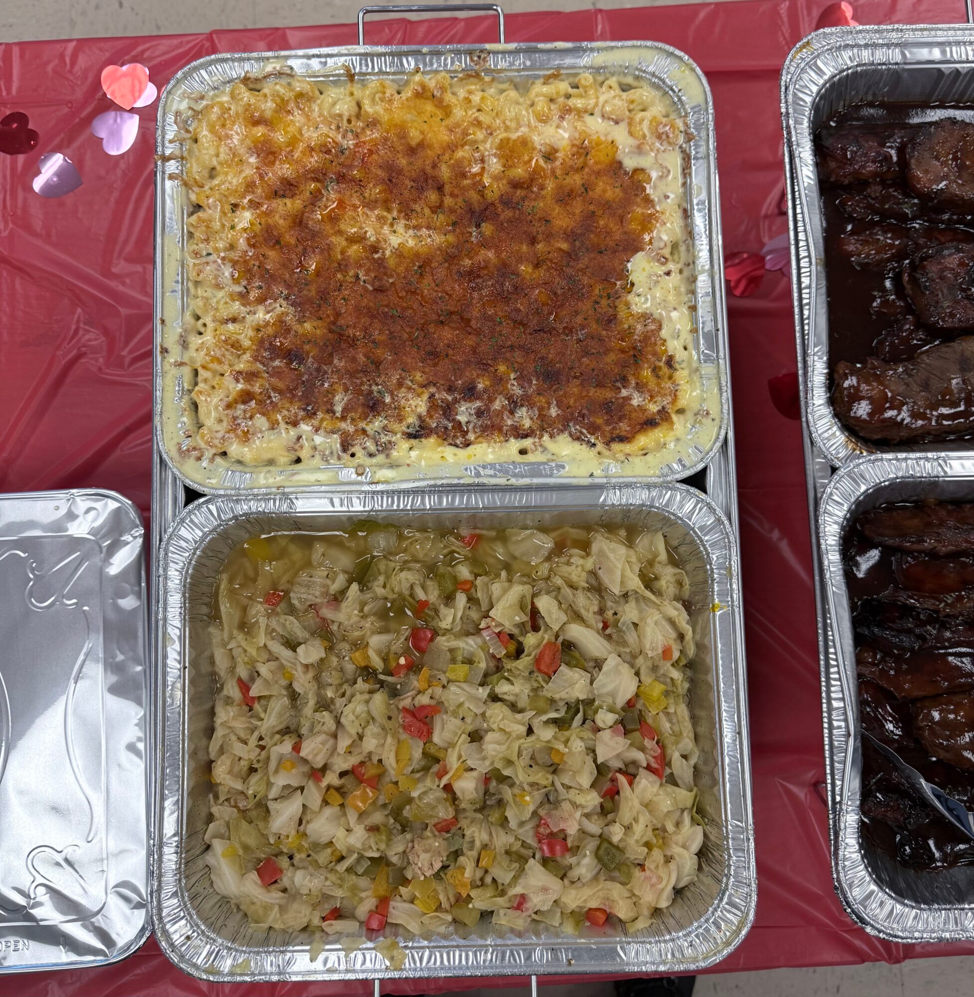 Two aluminum trays with baked dishes, one with a golden crust and the other with a vegetable mixture, on a red tablecloth.