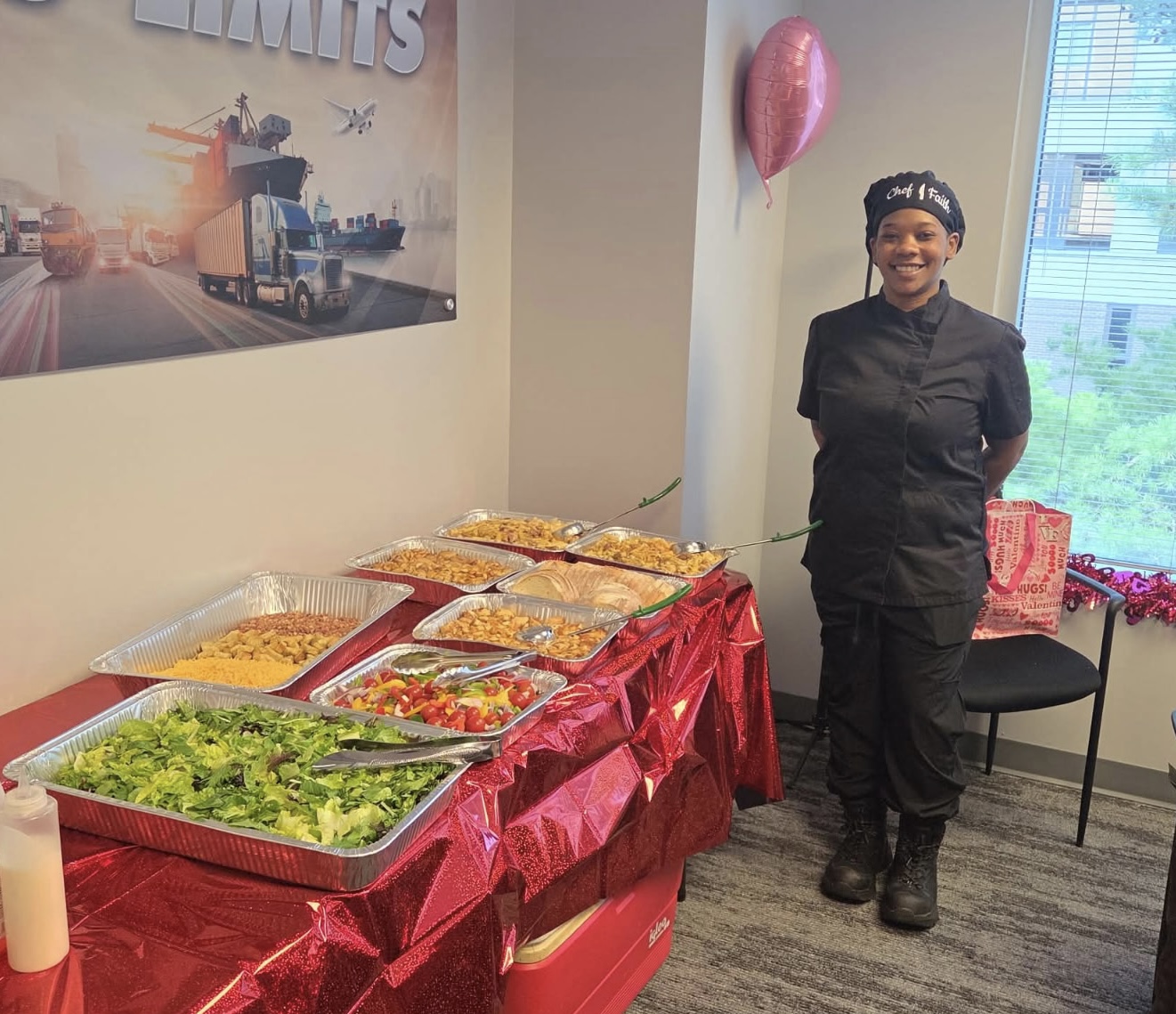 A woman in black uniform stands next to a table with salads and food trays, in a room with a window and pink balloon.