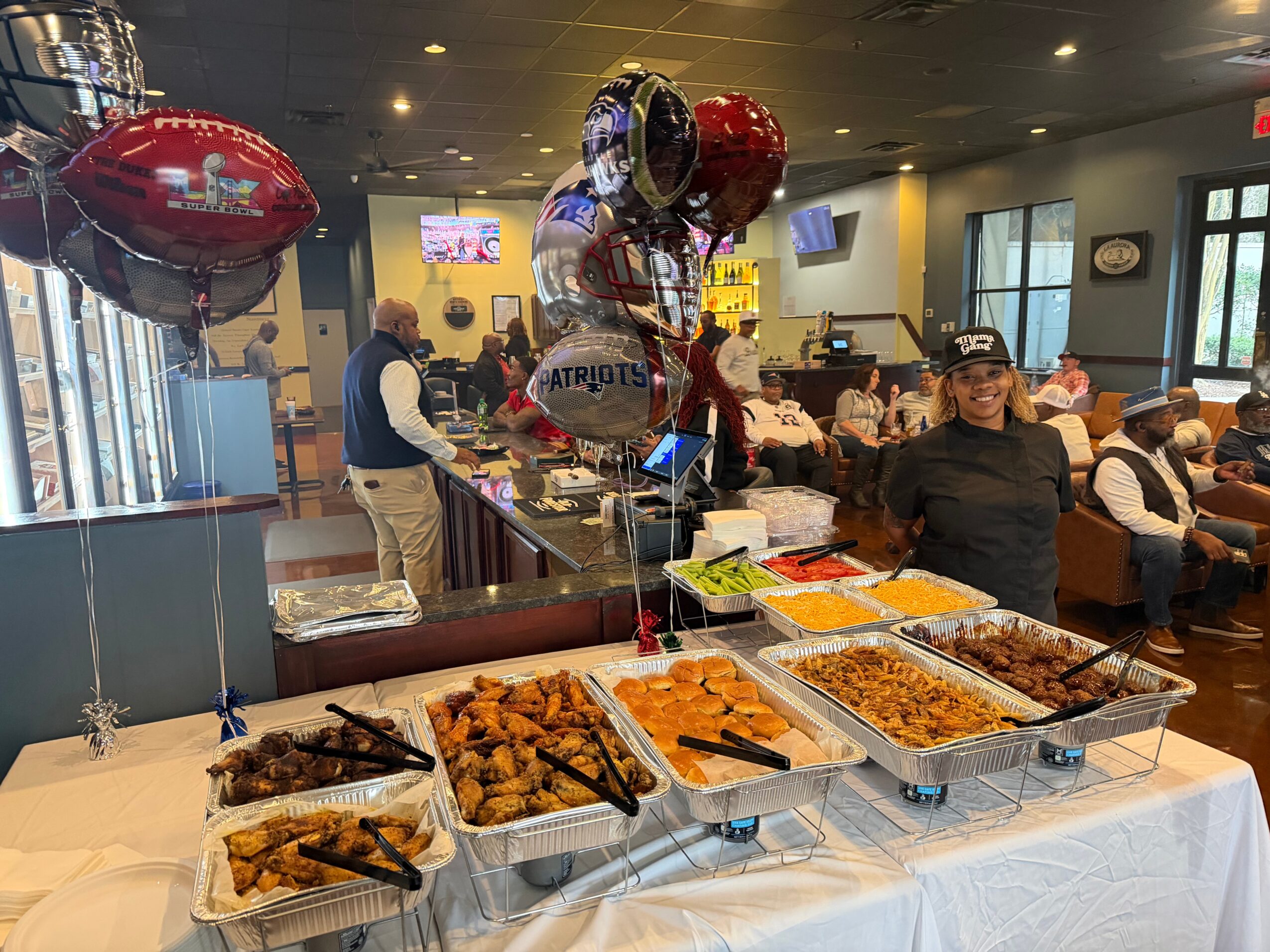 Buffet table with trays of fried chicken, ribs, and other dishes, balloons, and people in a restaurant setting.