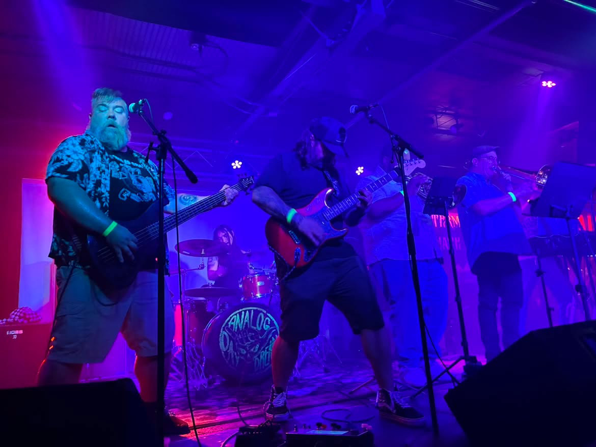 Band performing on stage with purple and blue lighting, four musicians playing guitars and drums, microphones in front, lively atmosphere.