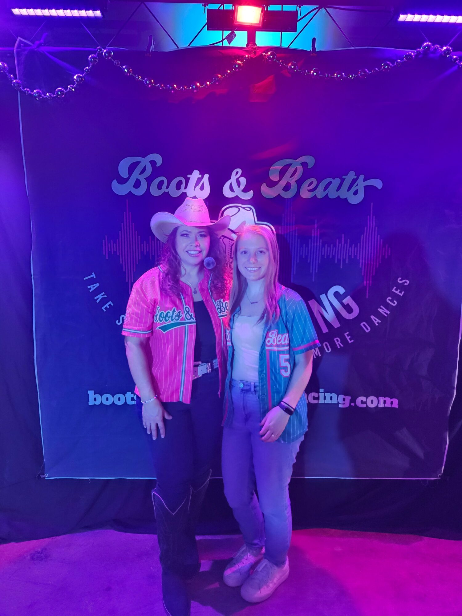 Two women stand together in front of a backdrop with text, smiling at the camera, in a dimly lit setting with purple lighting.