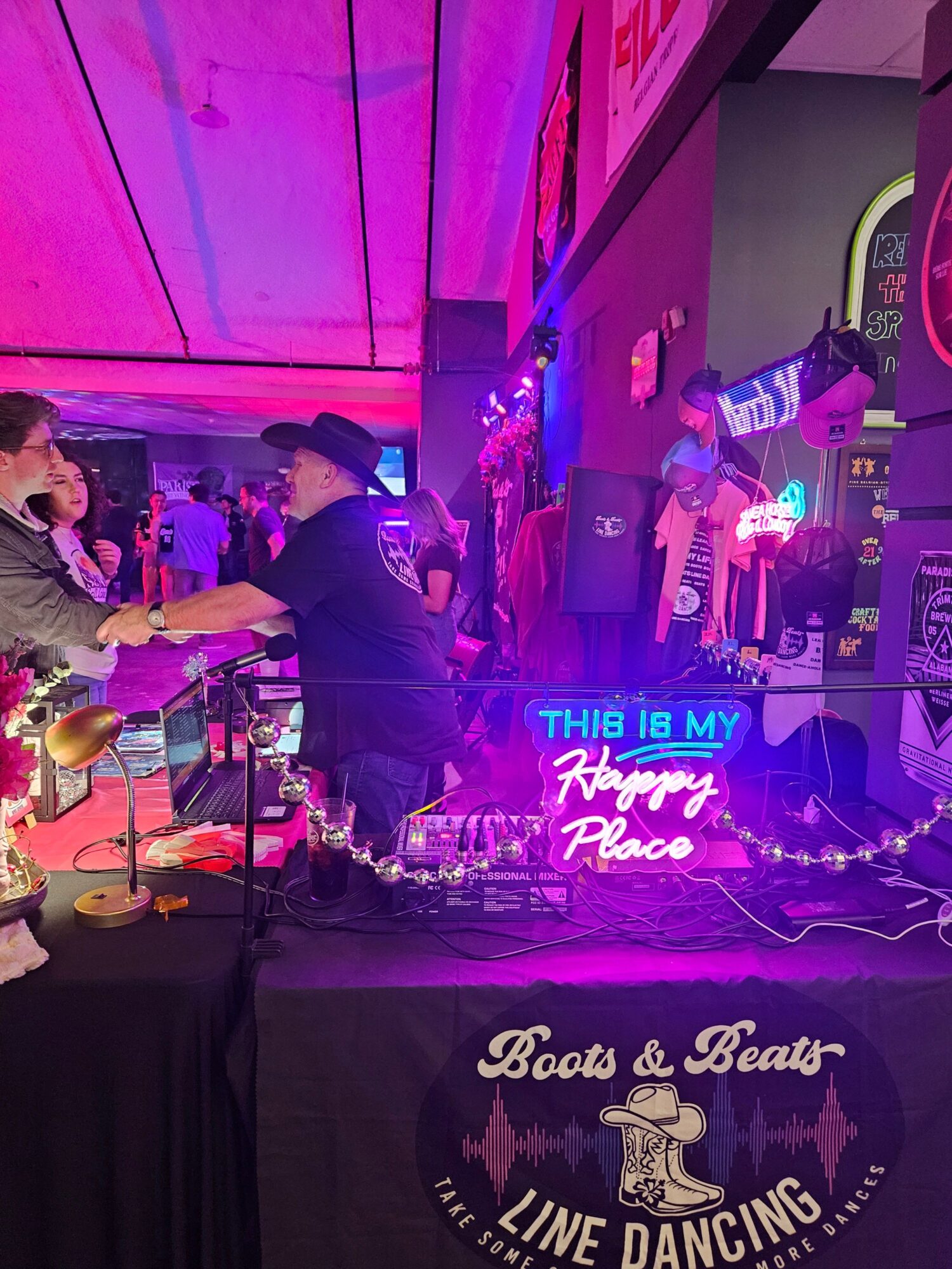 Person at a bar with purple lighting, standing near a table with a black tablecloth, neon signs, and a stage in the background.