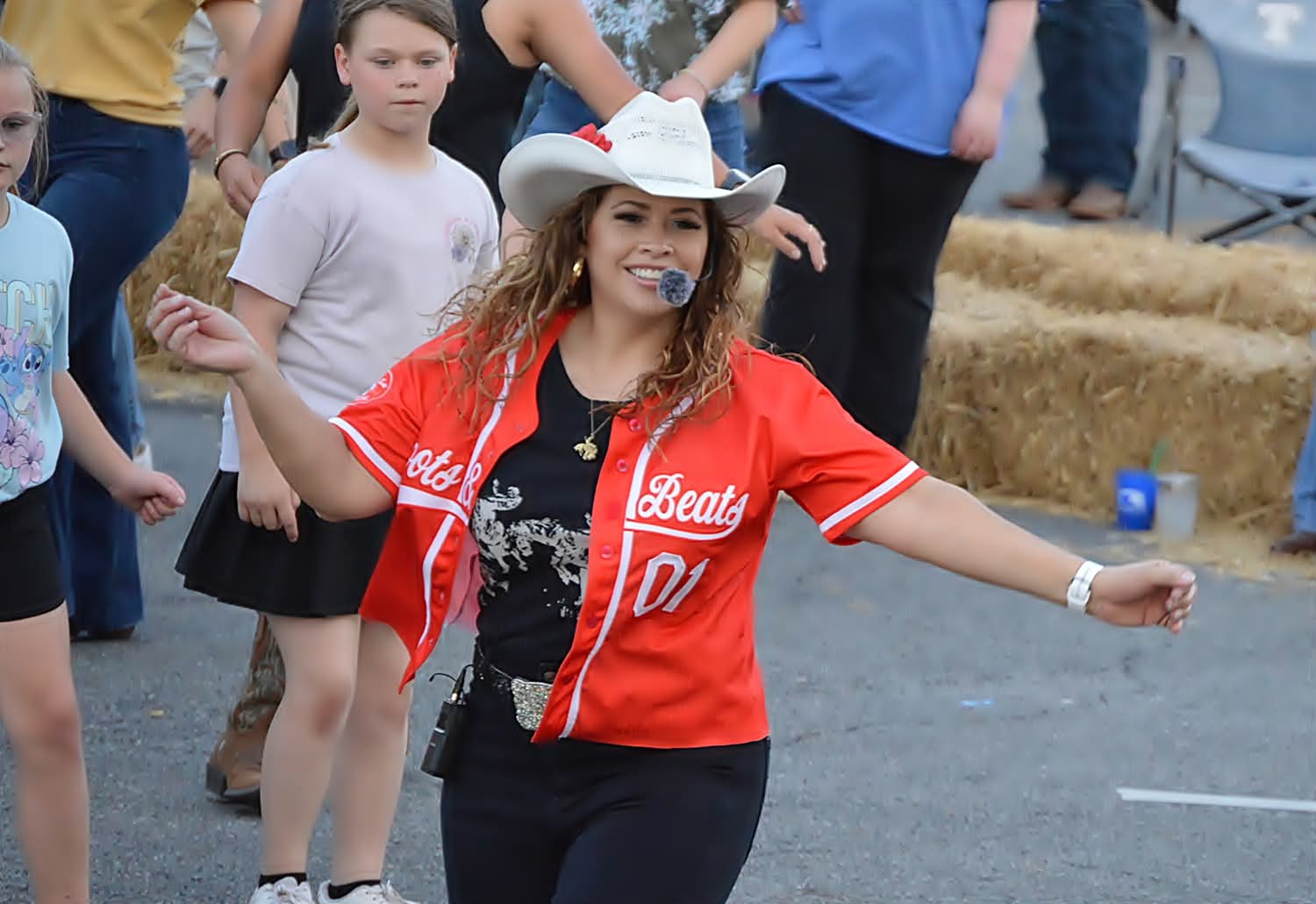 Young woman in a red baseball jersey and white hat smiling with arms outstretched at outdoor event.