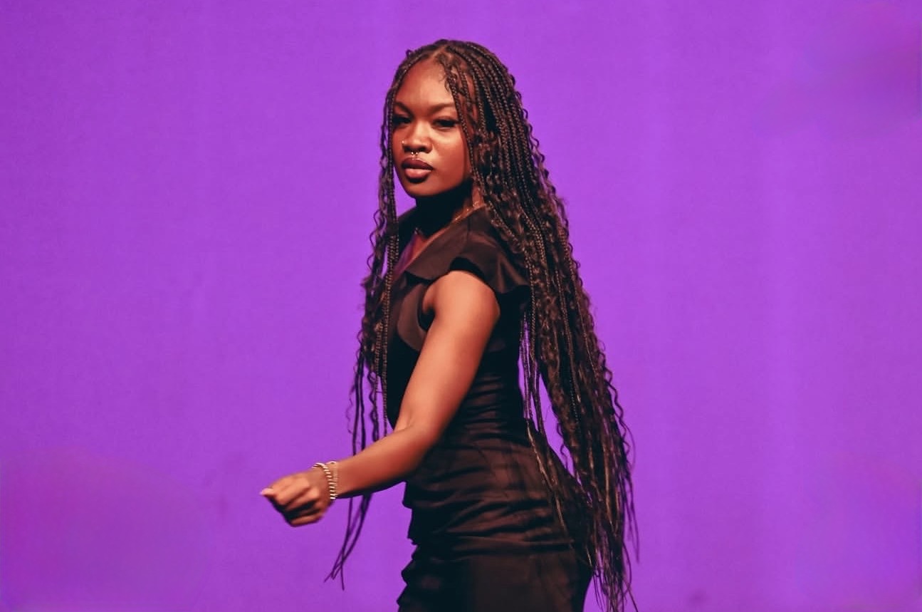 Young woman with long braided hair stands against a purple background, wearing a black outfit, looking at the camera.