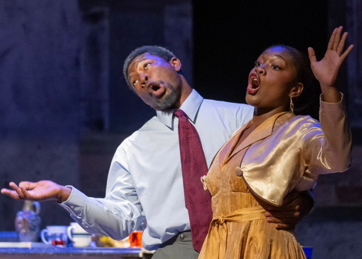 Two performers singing on stage, one with a white shirt and red tie, the other in a gold dress, expressive gestures.