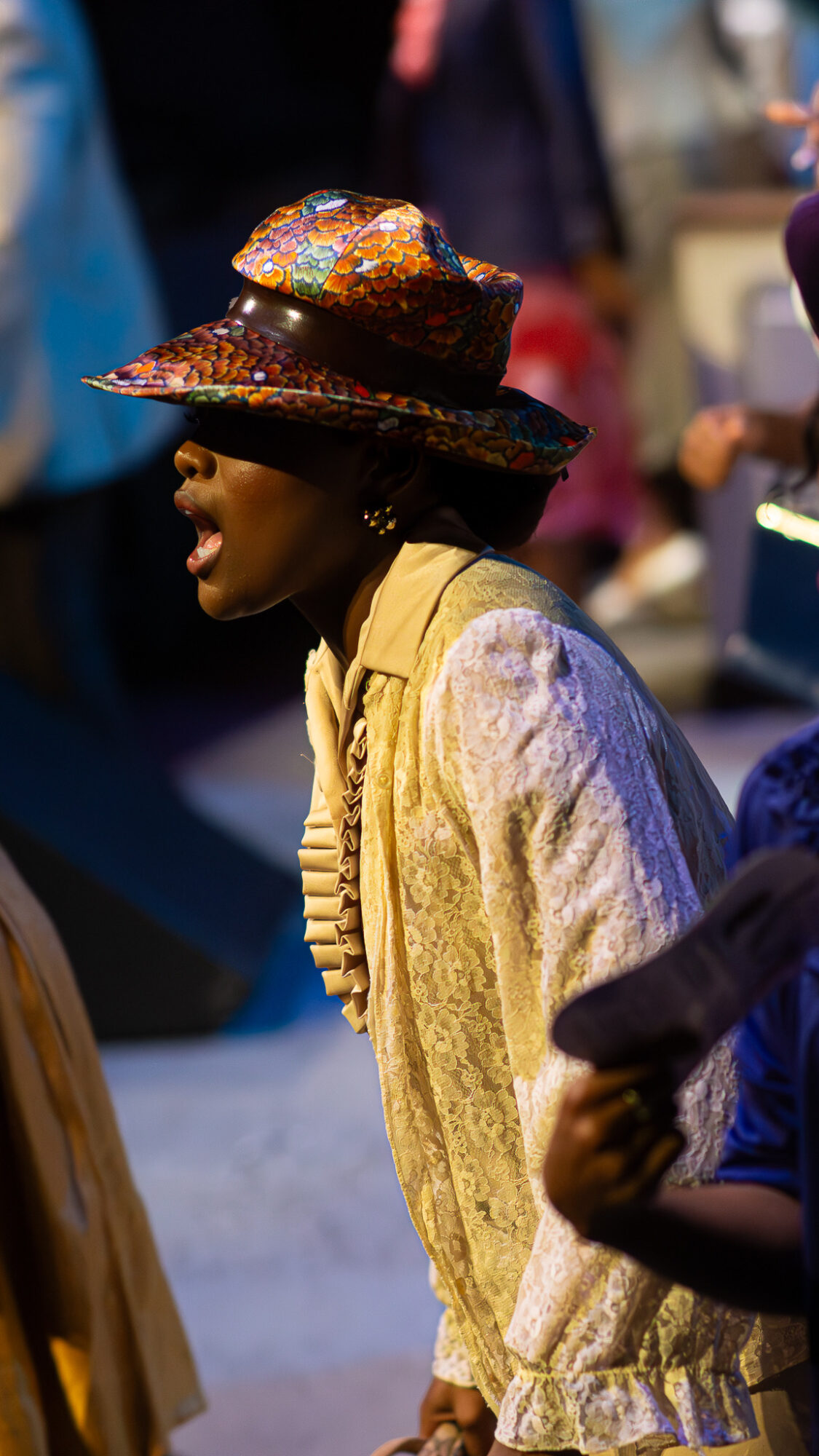 Person wearing a colorful, wide-brimmed hat and beige jacket, standing in a crowded indoor setting.