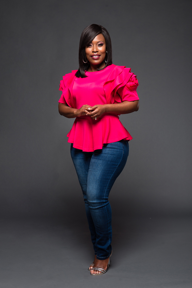 Woman in pink top and jeans standing against gray background, smiling, hands clasped in front.