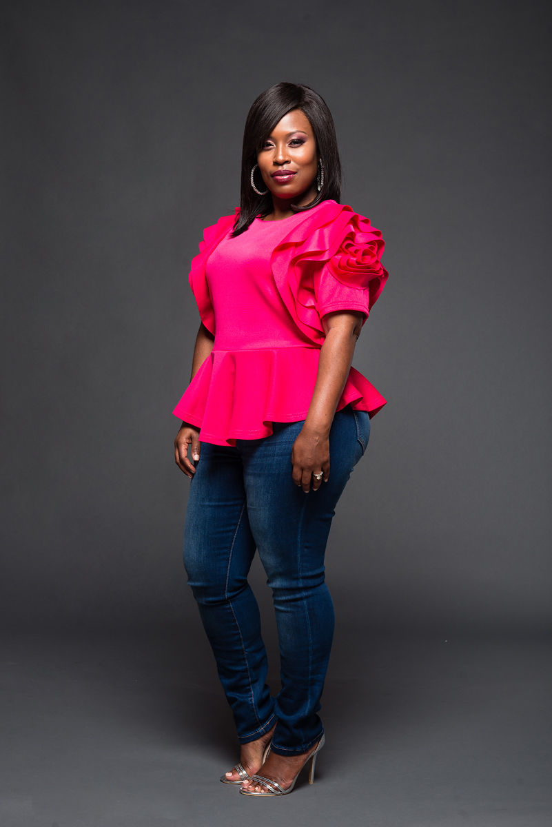 Woman in bright pink top with ruffled sleeves and blue jeans, standing against a dark background.