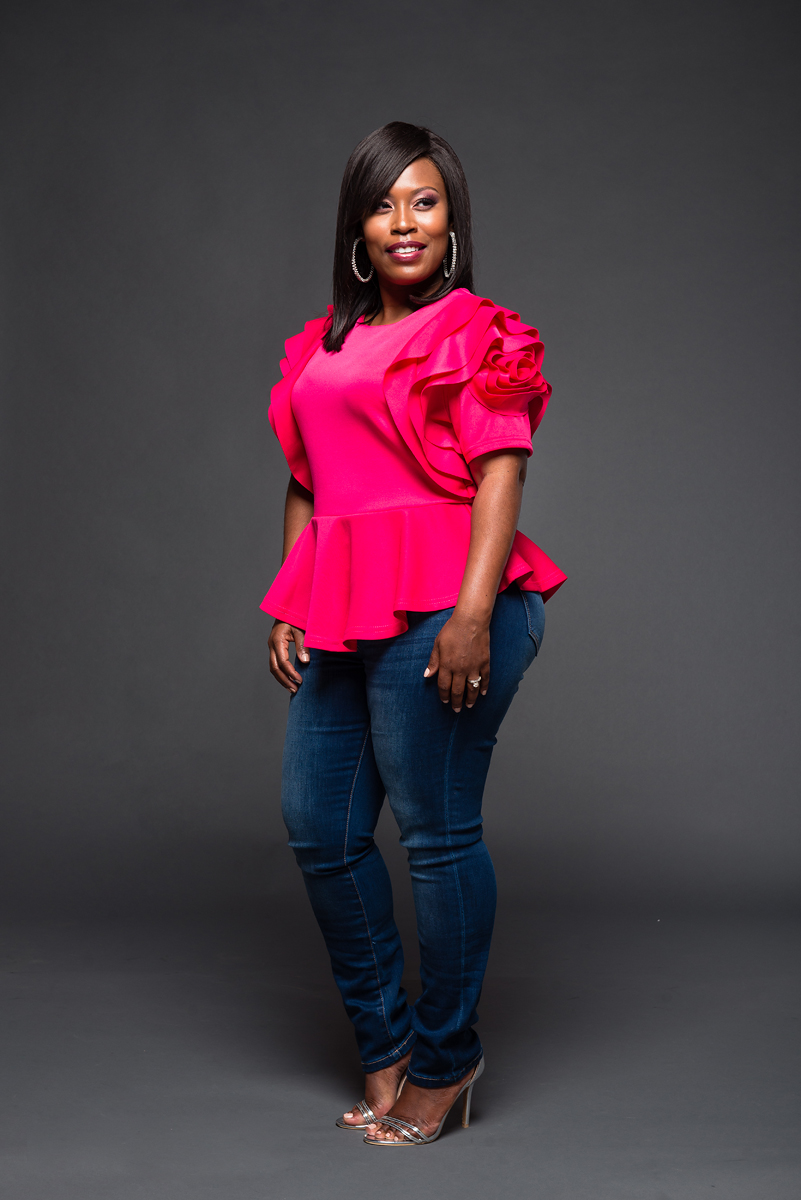 Woman in pink top and jeans standing against a dark background, smiling, with hands at sides, wearing heels.