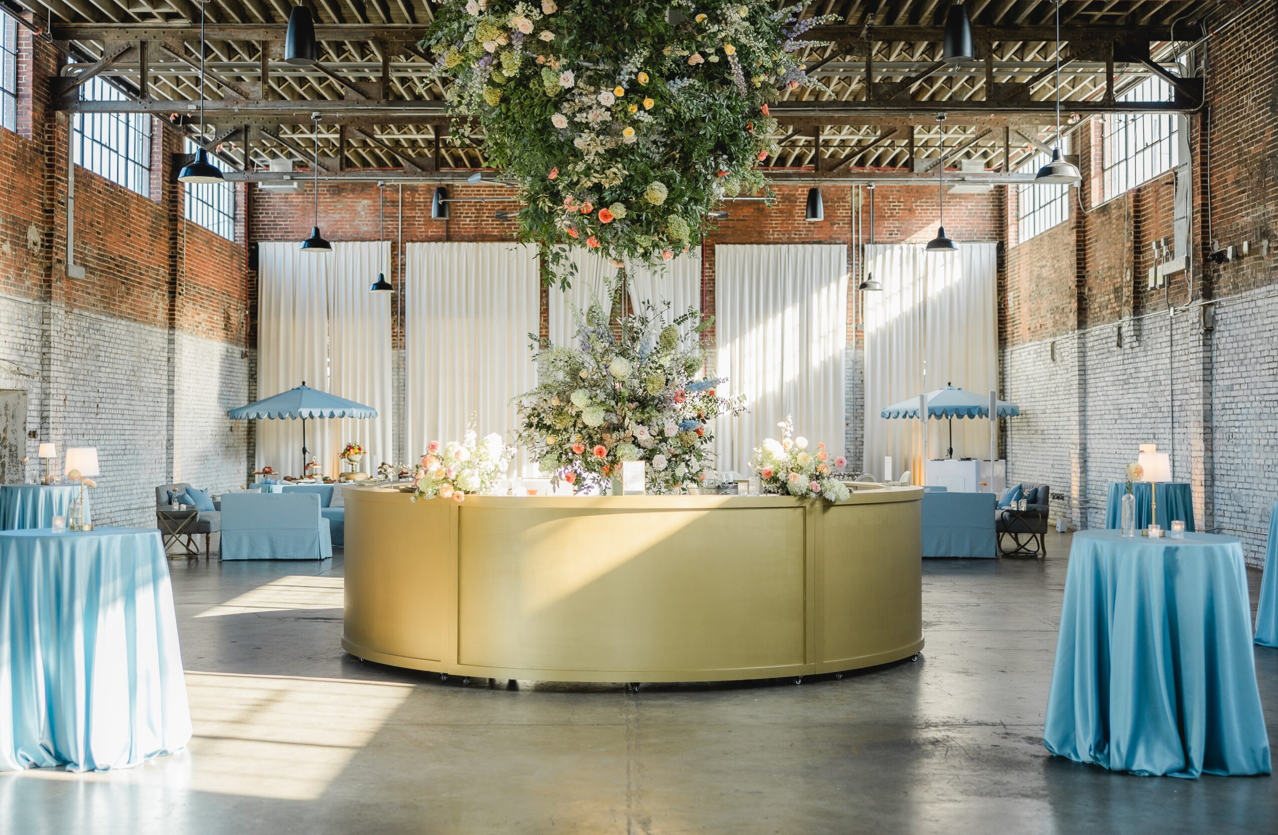Event space with a large floral arrangement, round bar, and tables with blue tablecloths, brick walls, and high ceilings.