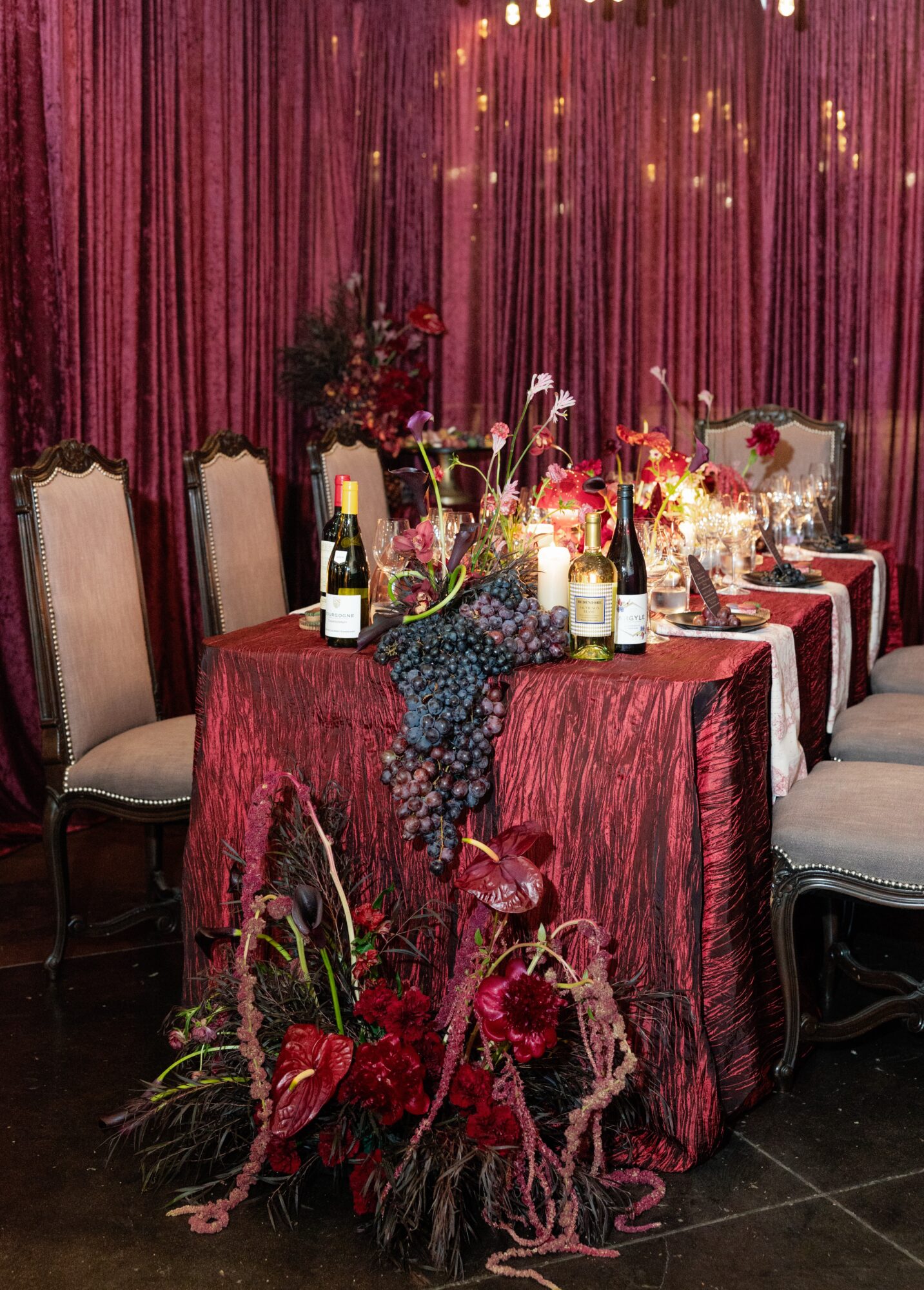 Elegant dining table with red tablecloth, floral centerpiece, candles, wine bottles, and chairs, set against a pink curtain backdrop.
