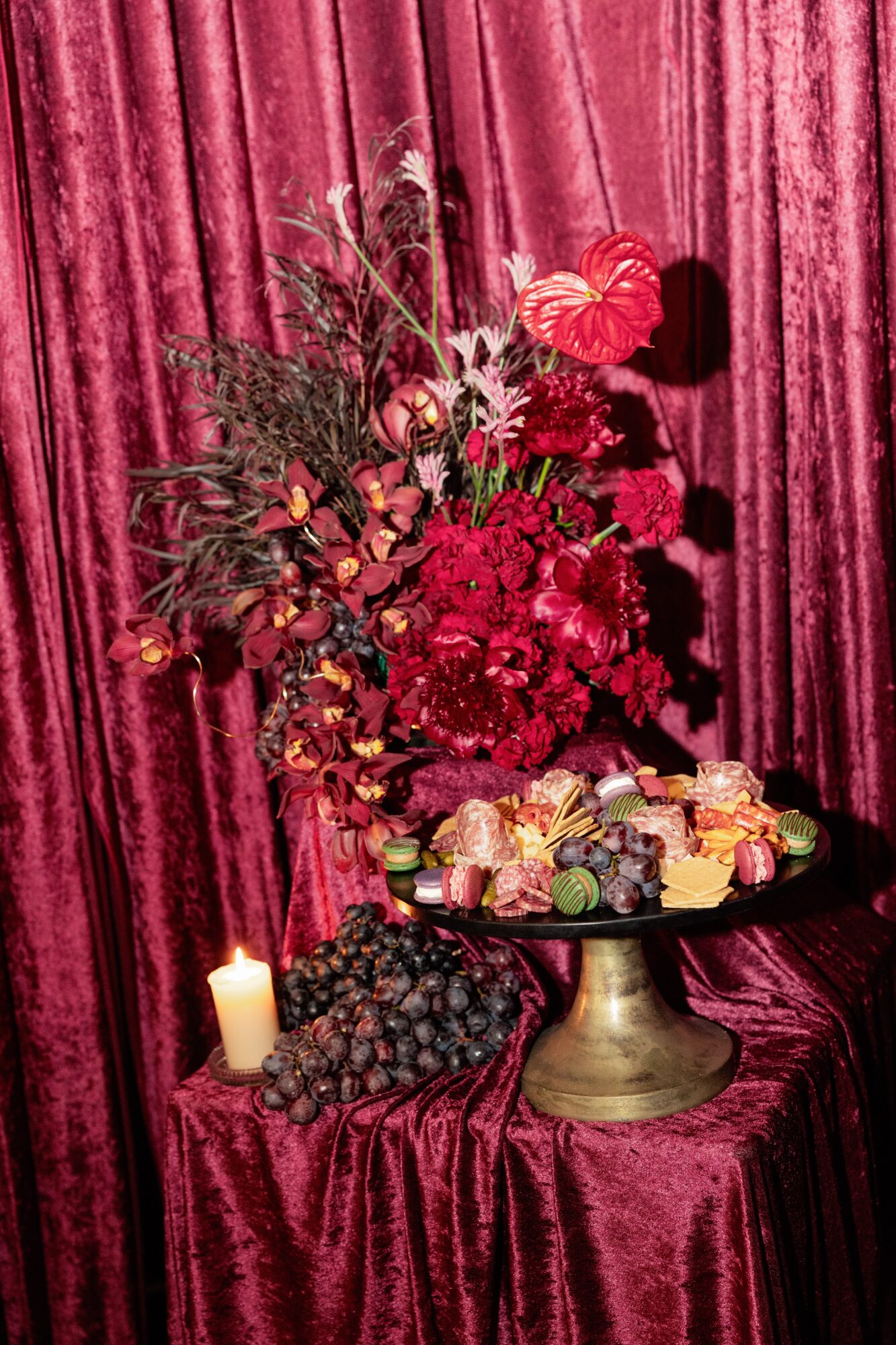 Flower arrangement with red and pink flowers on a gold stand, surrounded by grapes, candies, and a candle, against a red curtain.