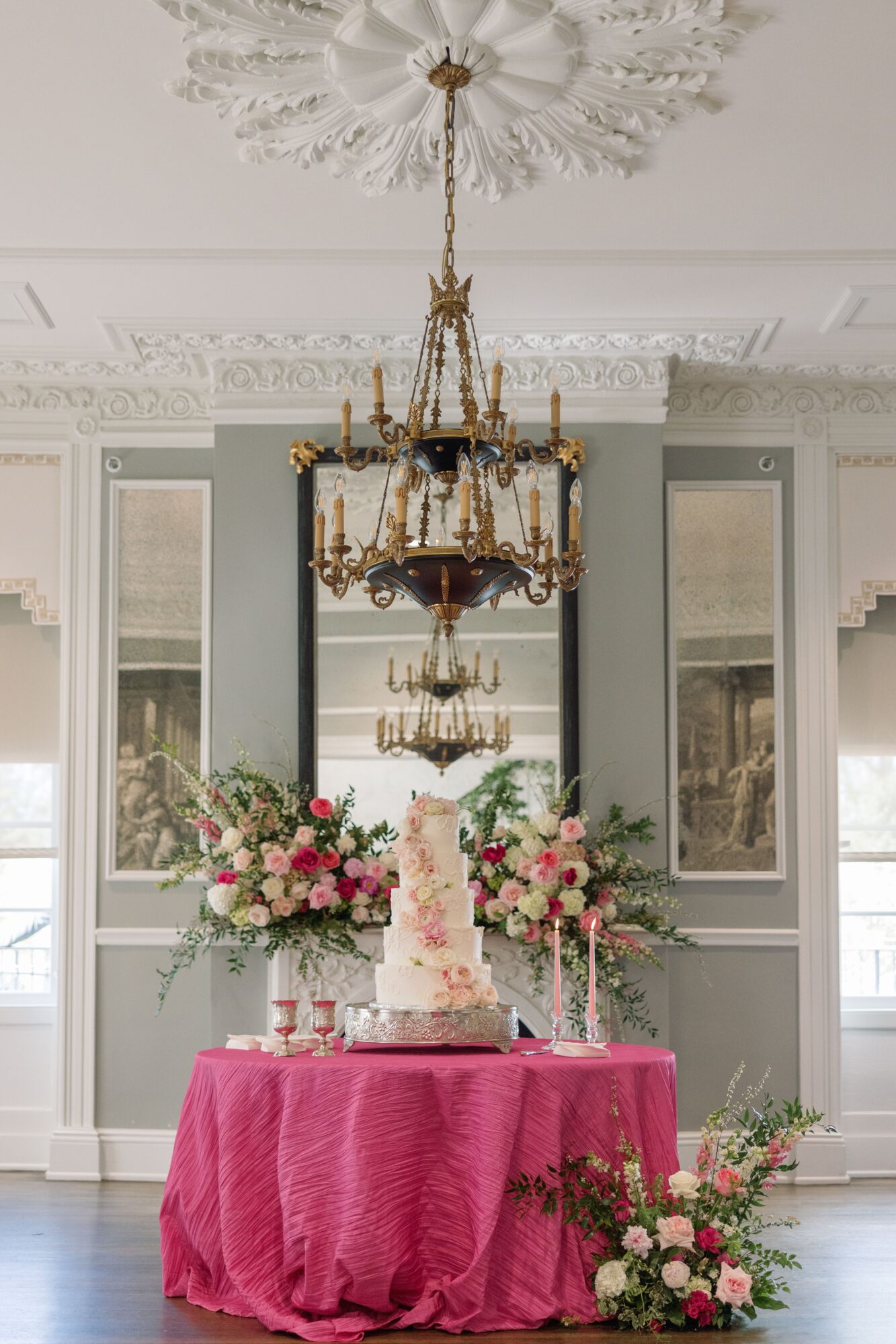 Elegant room with chandelier, decorated table with pink tablecloth, floral arrangements, and a tiered cake, windows in background.