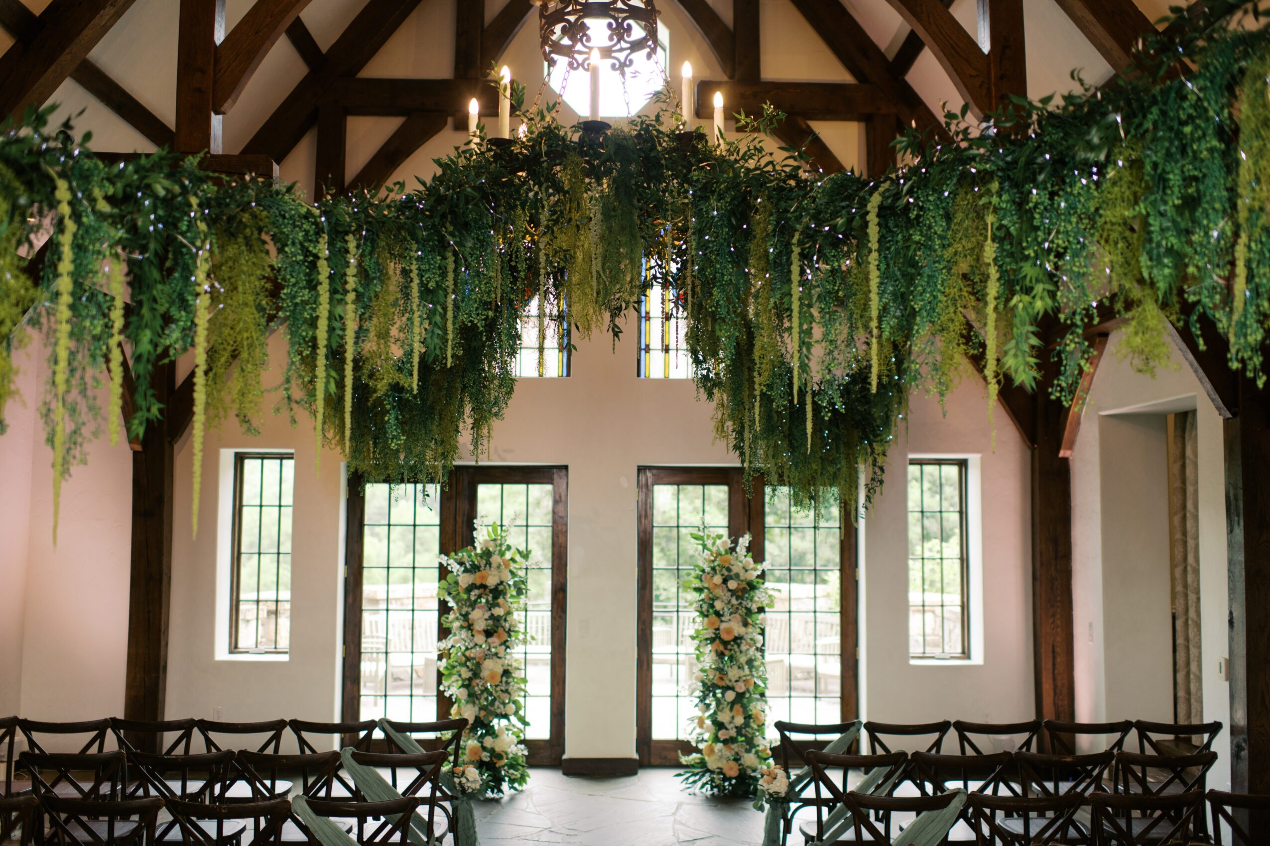 Interior of a decorated wedding or event space with hanging greenery, floral arrangements, and chairs arranged for guests.