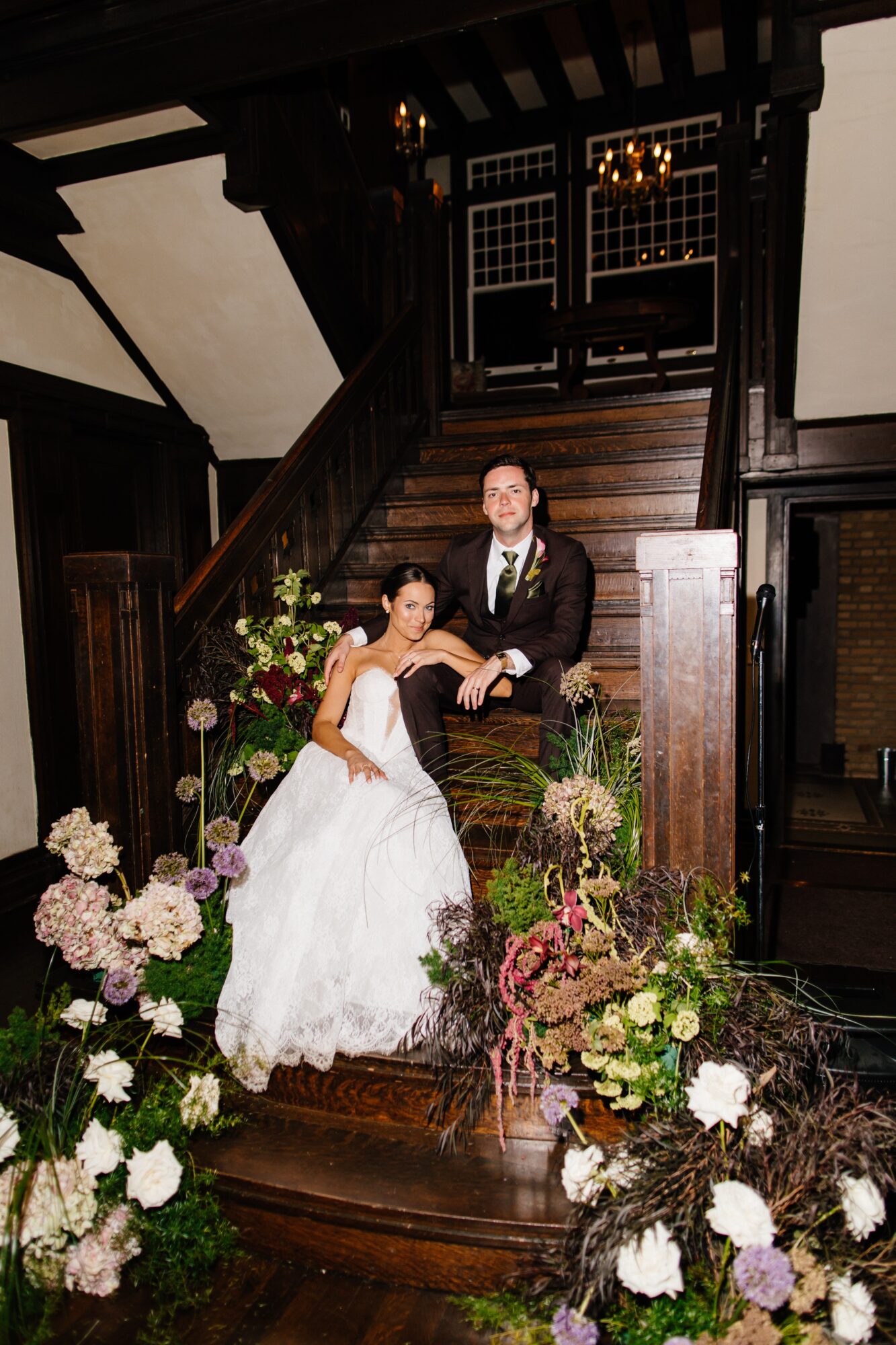 Bride and groom sitting on staircase with floral arrangements, dark wood interior, and staircase behind them.