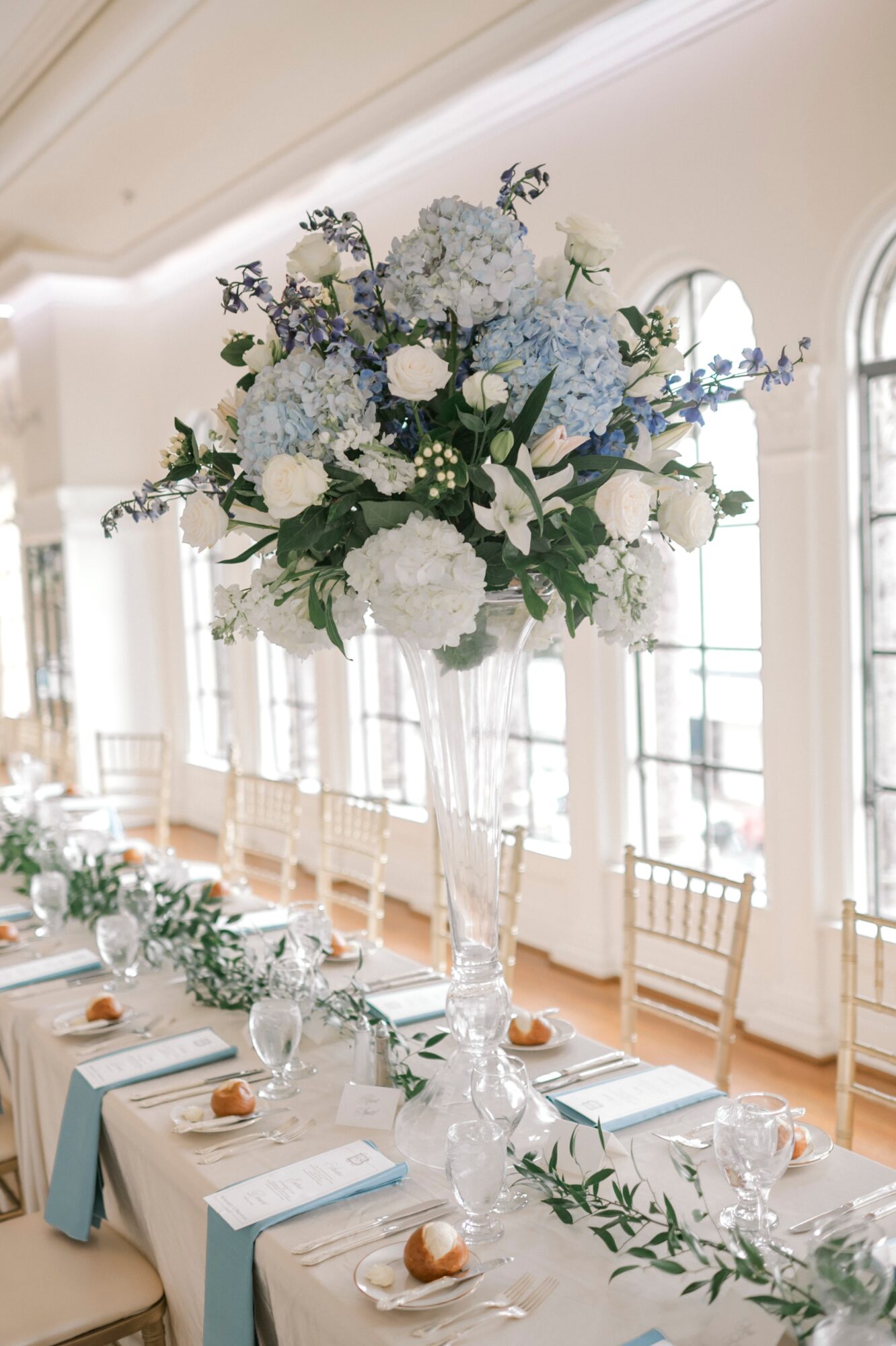 Elegant table centerpiece with blue and white flowers in a tall glass vase, set in a bright room with large windows.