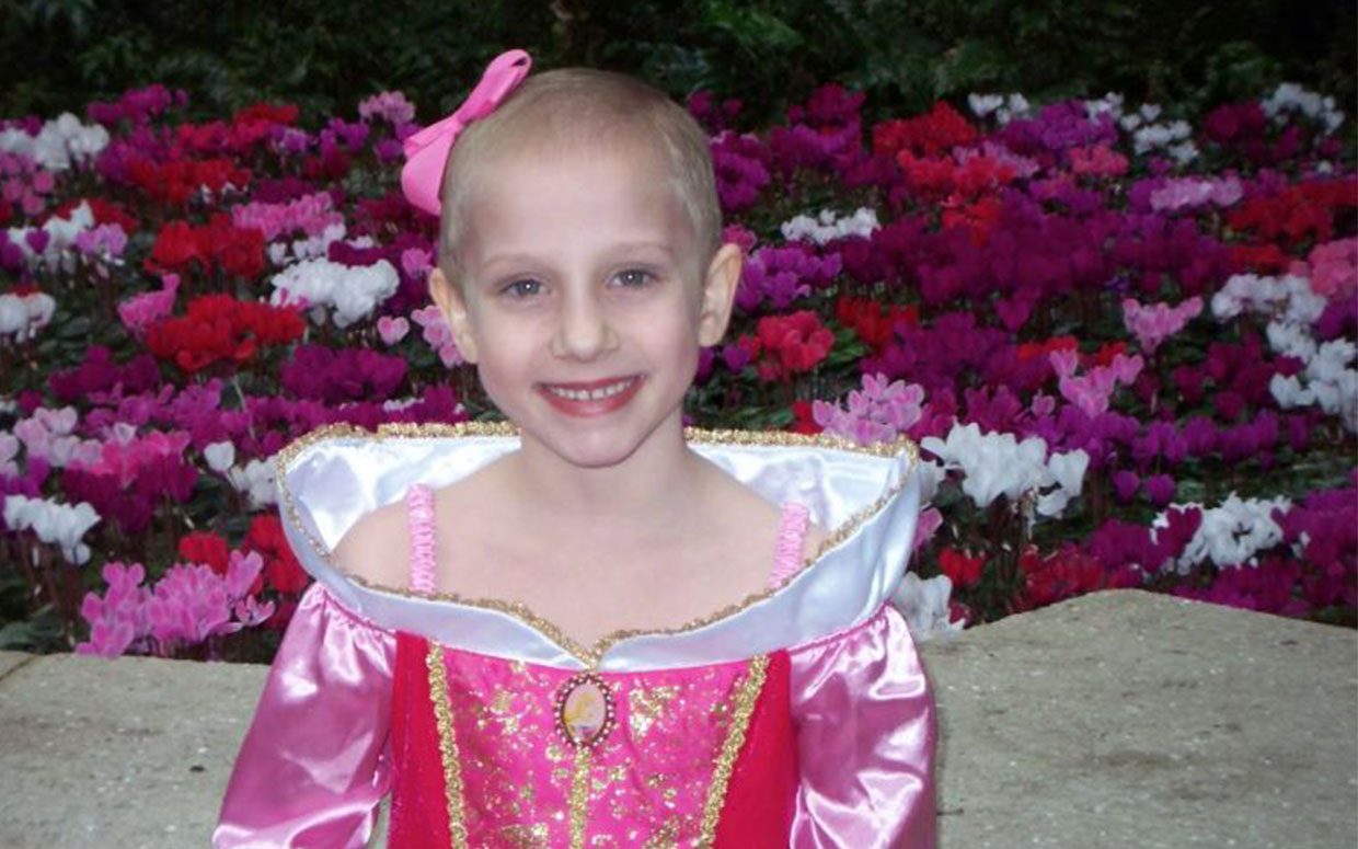 Young girl in a pink princess dress smiling in front of colorful flowers, outdoors.