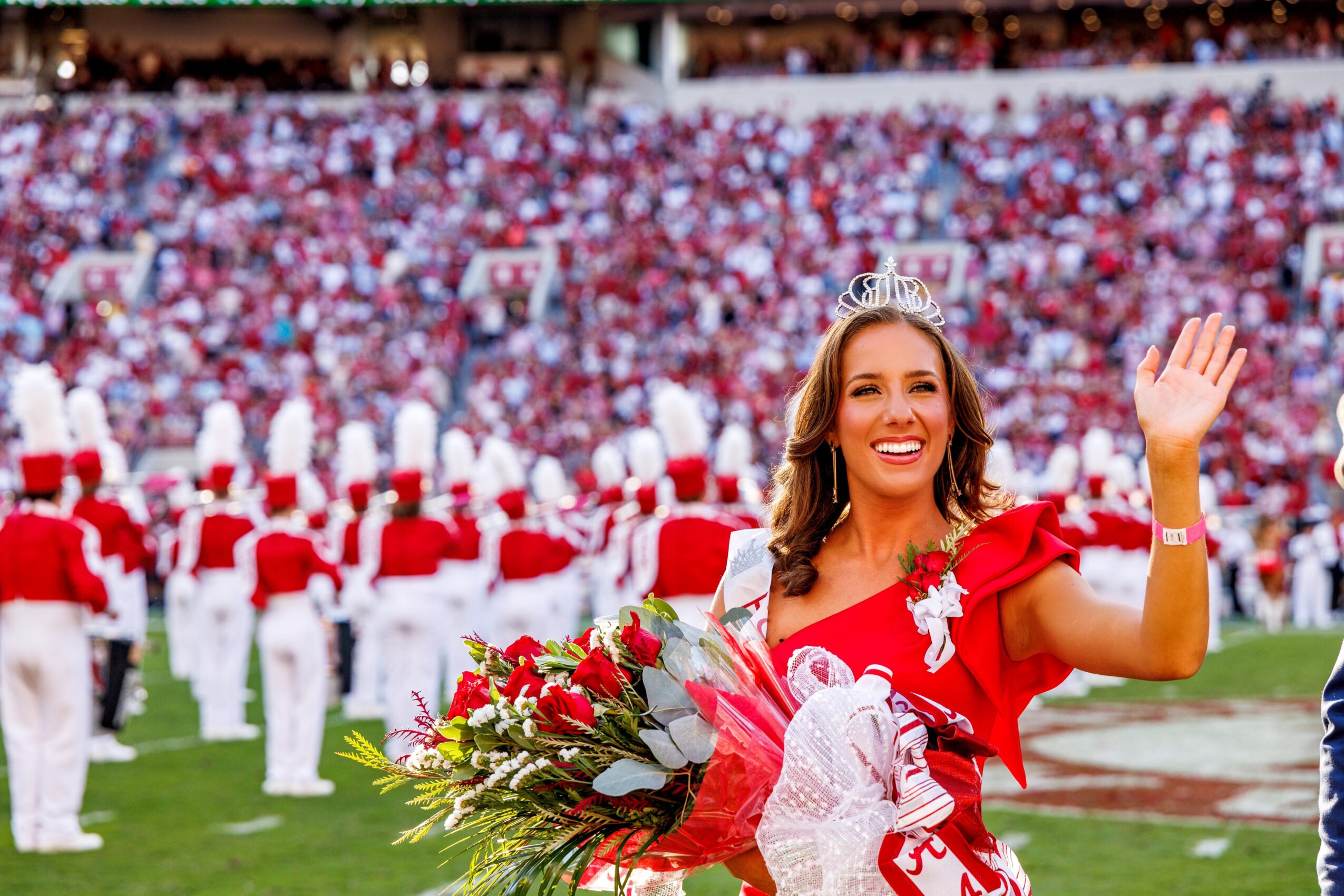 Woman in red dress holding a bouquet, waving, in a stadium with a large crowd and marching band in the background.