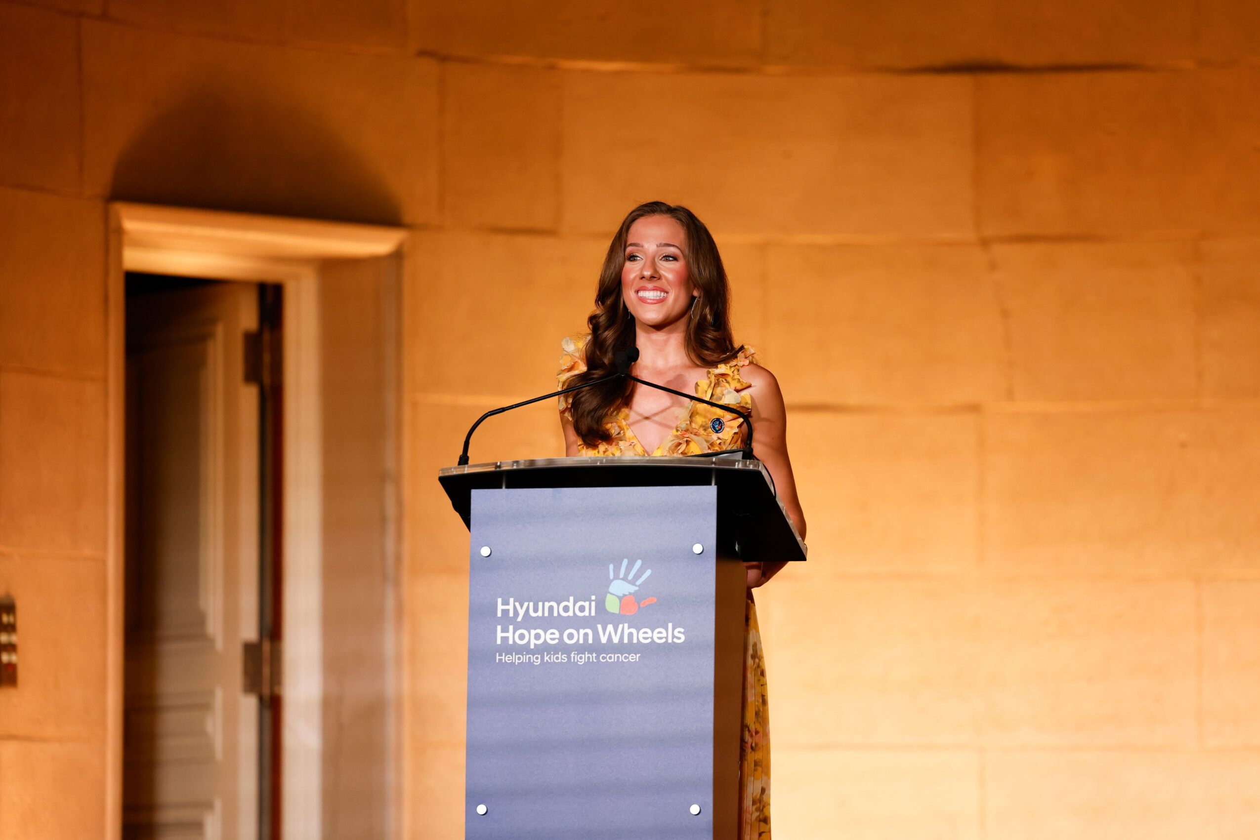 Woman standing at podium giving speech, smiling, in a warm-colored room.