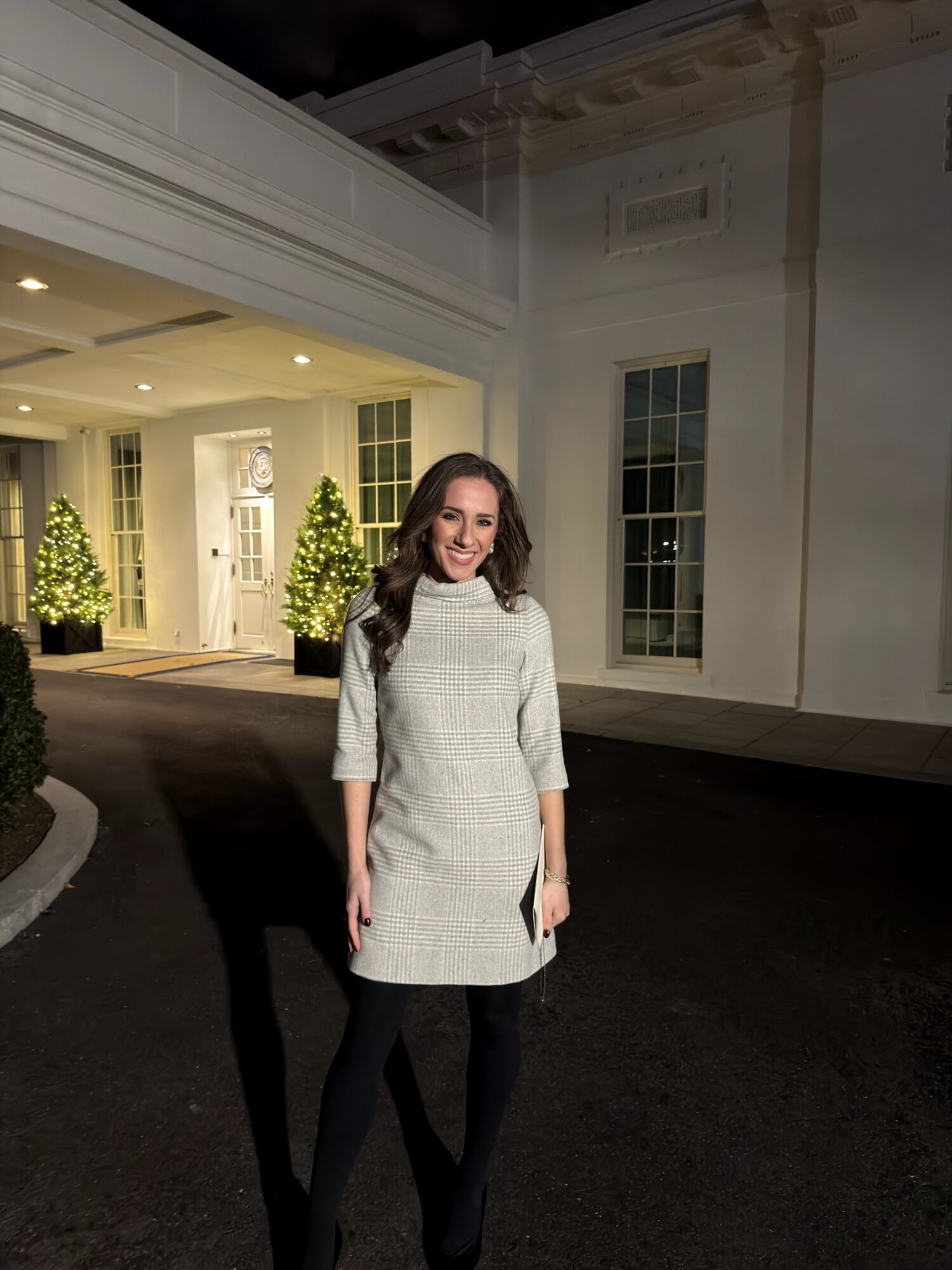 Woman standing outside a building at night, smiling, wearing a light-colored dress and black leggings, with lit windows and decorated trees in background.