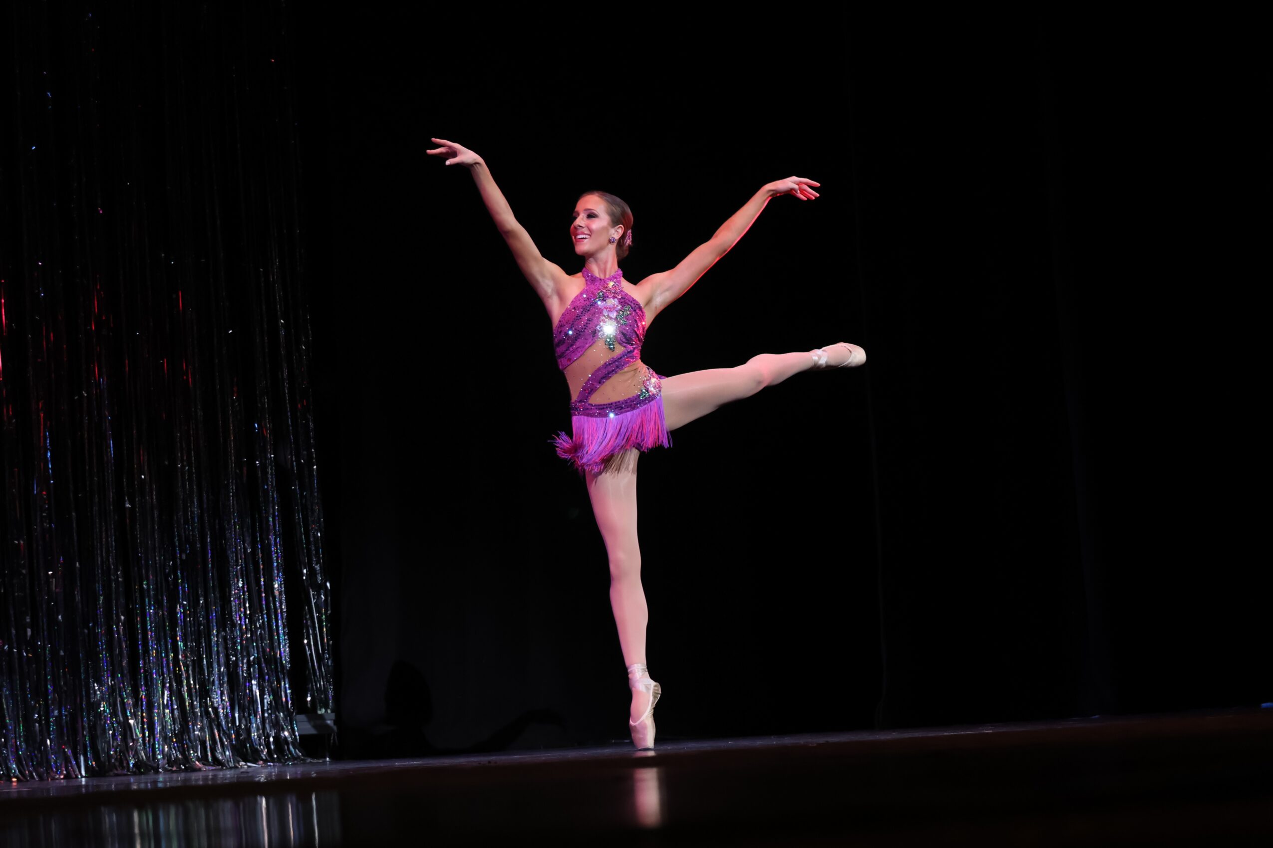 Ballet dancer in purple costume performing a pose on stage with a black background.